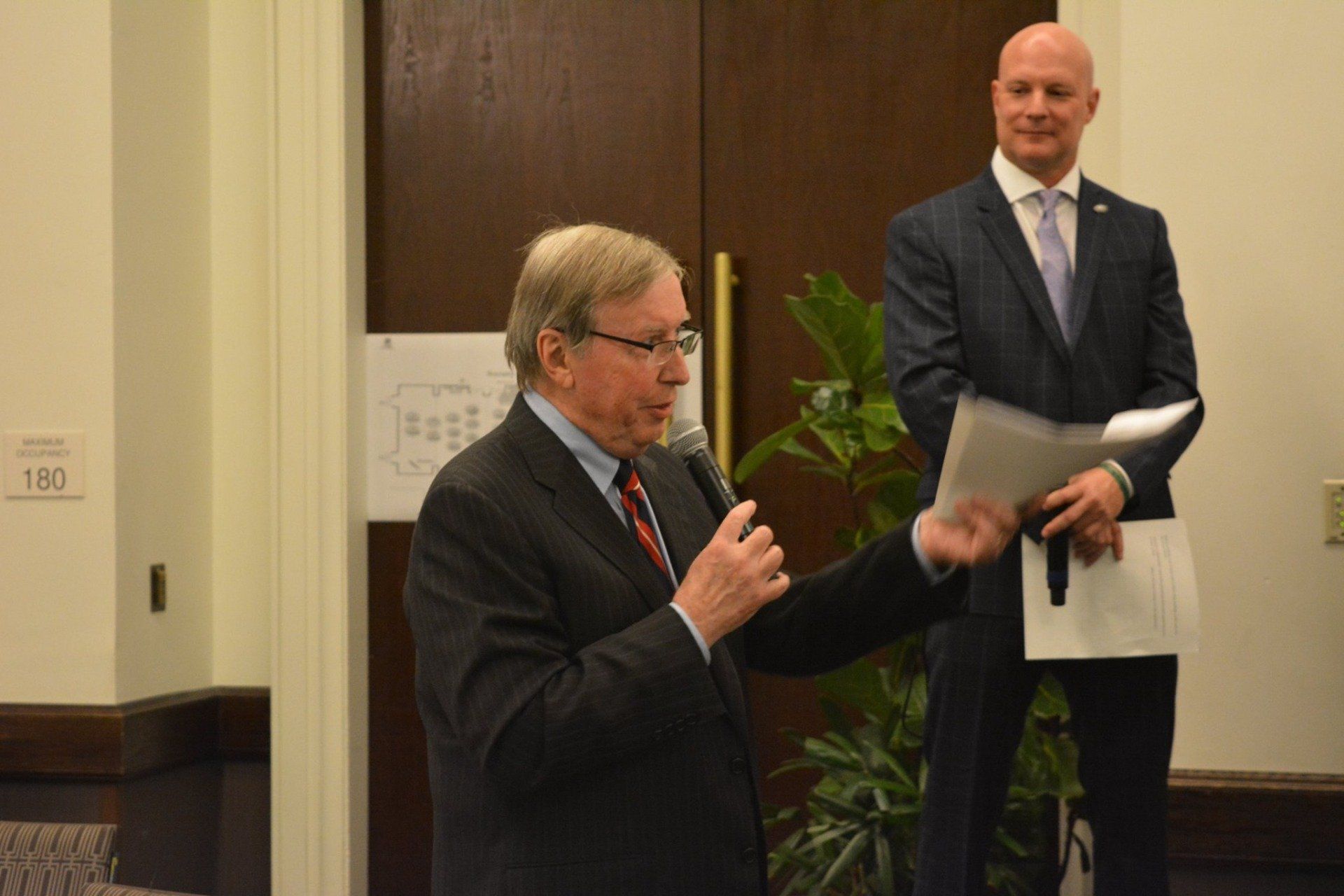 A man in a suit is speaking into a microphone while another man holds a piece of paper.