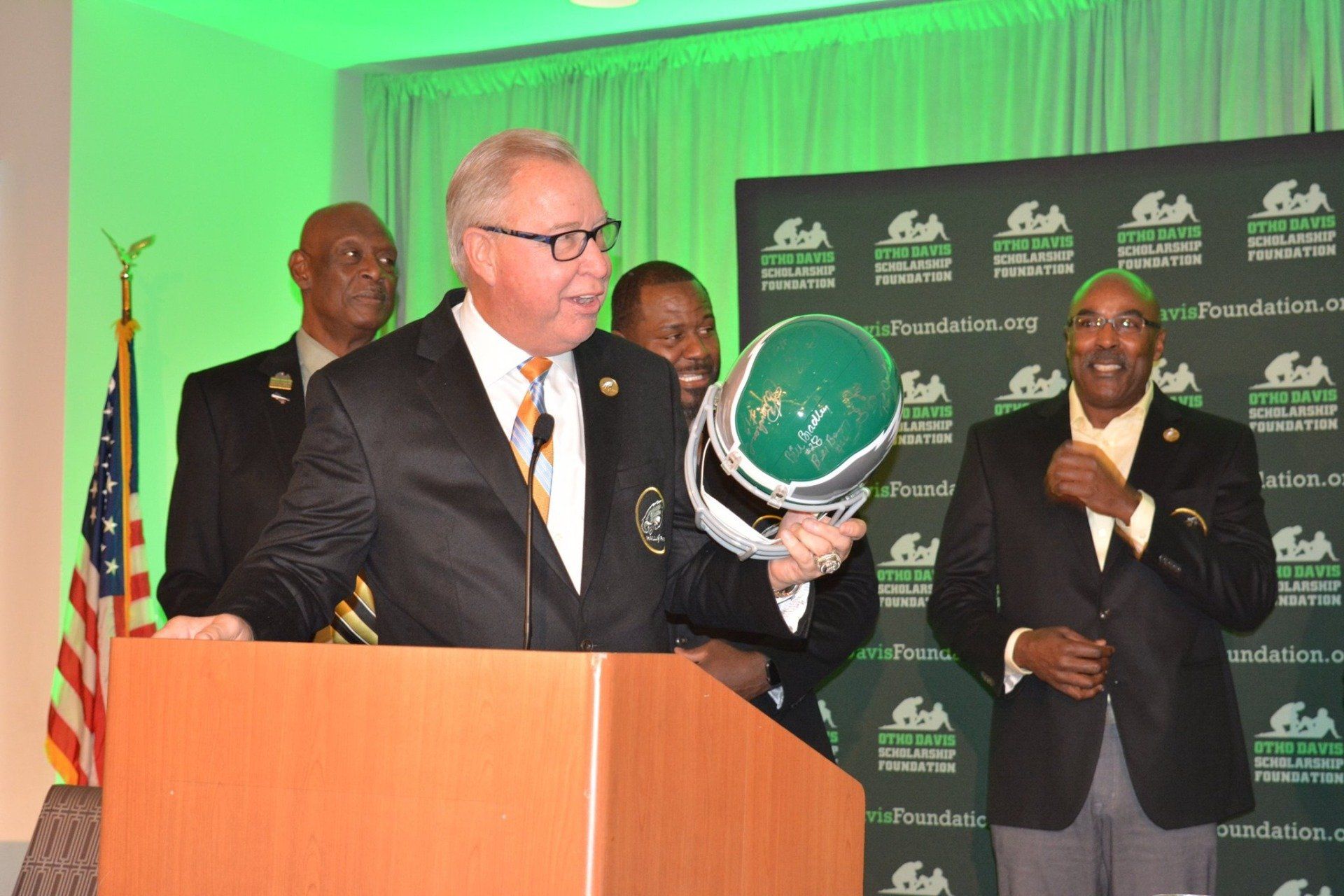 A man in a suit stands at a podium holding a helmet