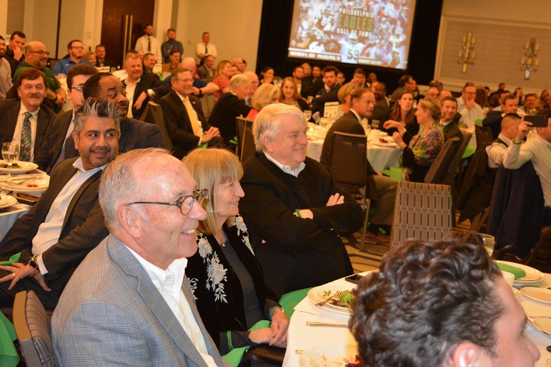 A group of people are sitting at tables in front of a large screen.