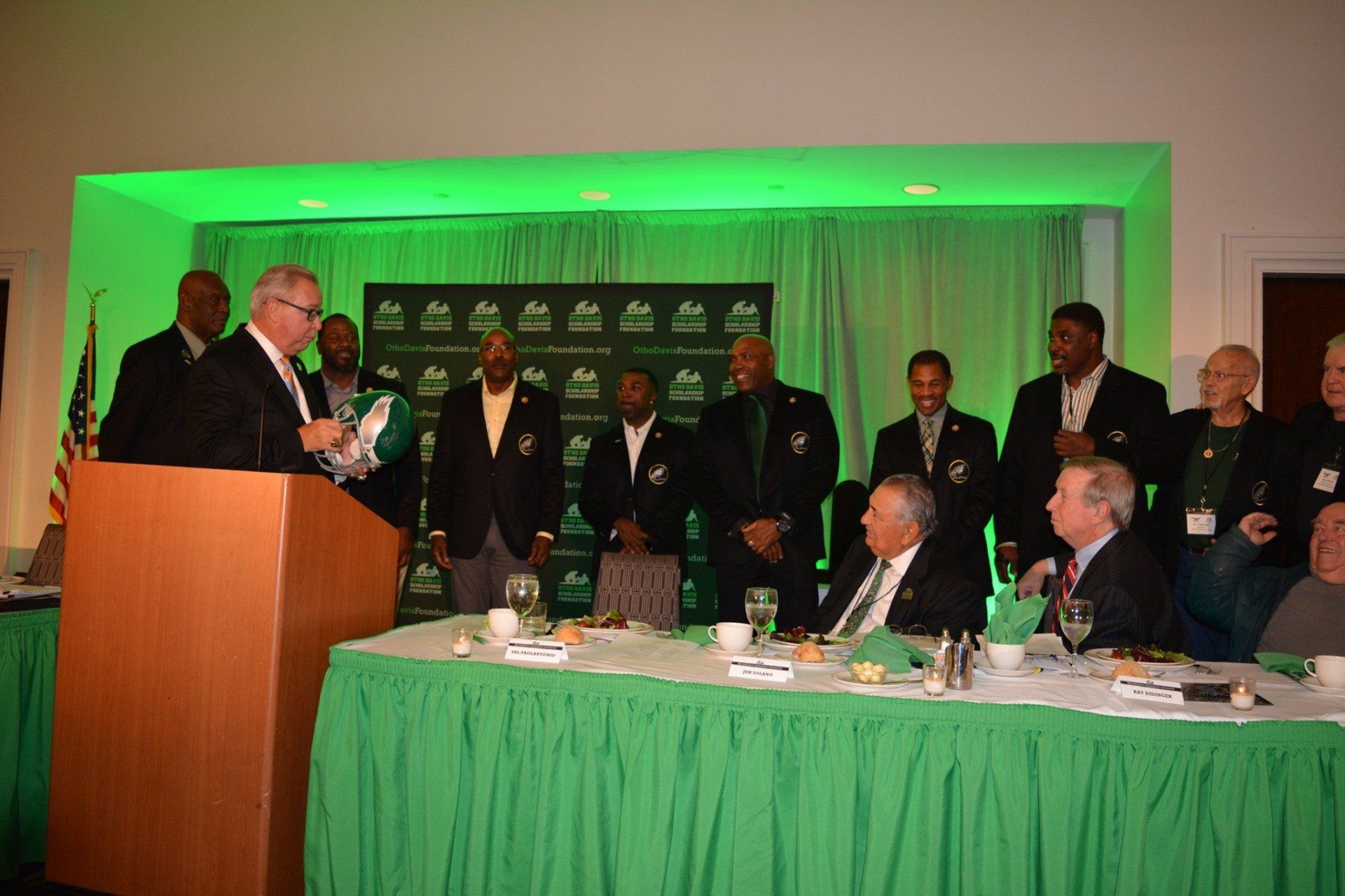 A man is giving a speech at a podium in front of a group of men sitting at a table.
