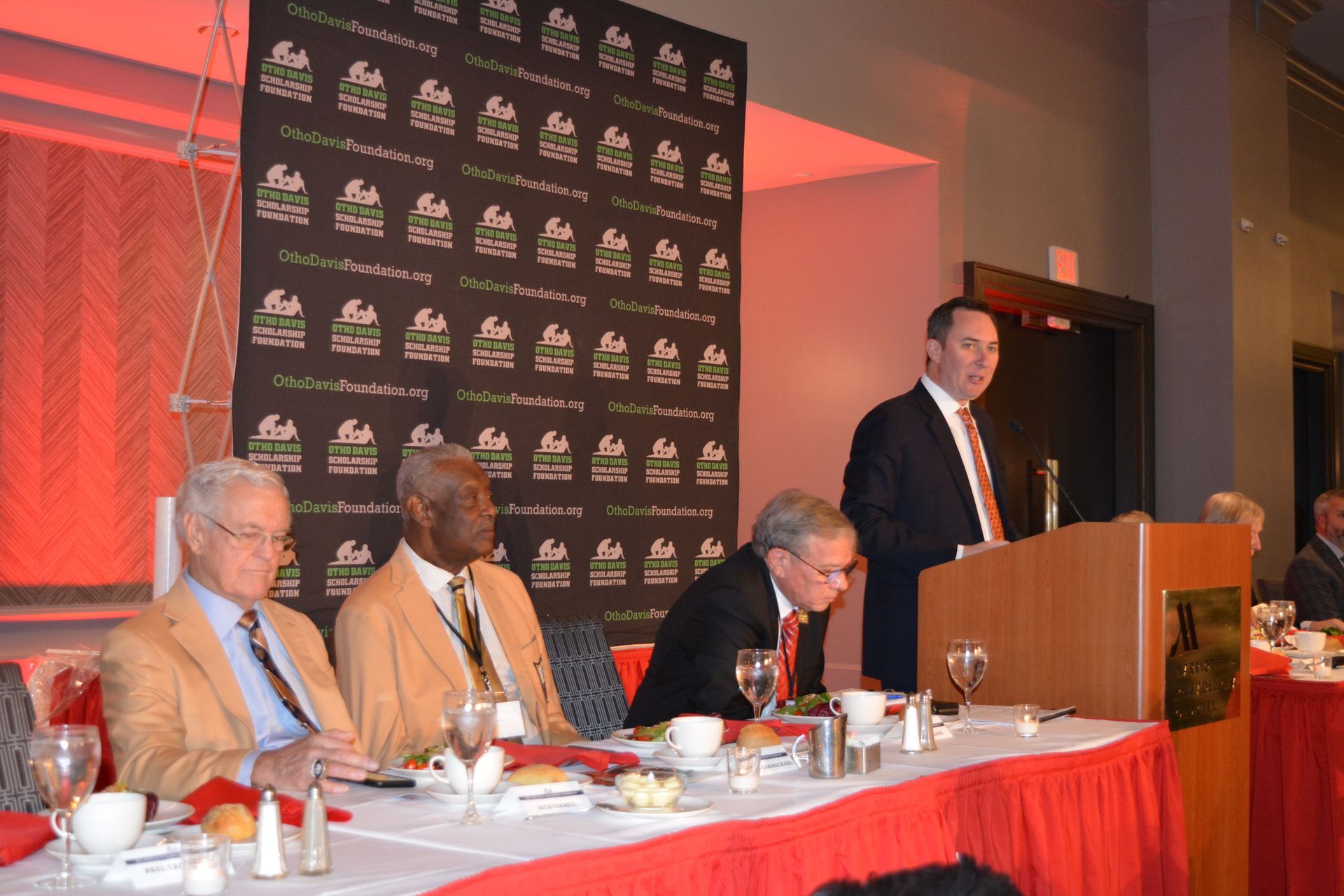 A group of men are sitting at a table with a man standing at a podium giving a speech