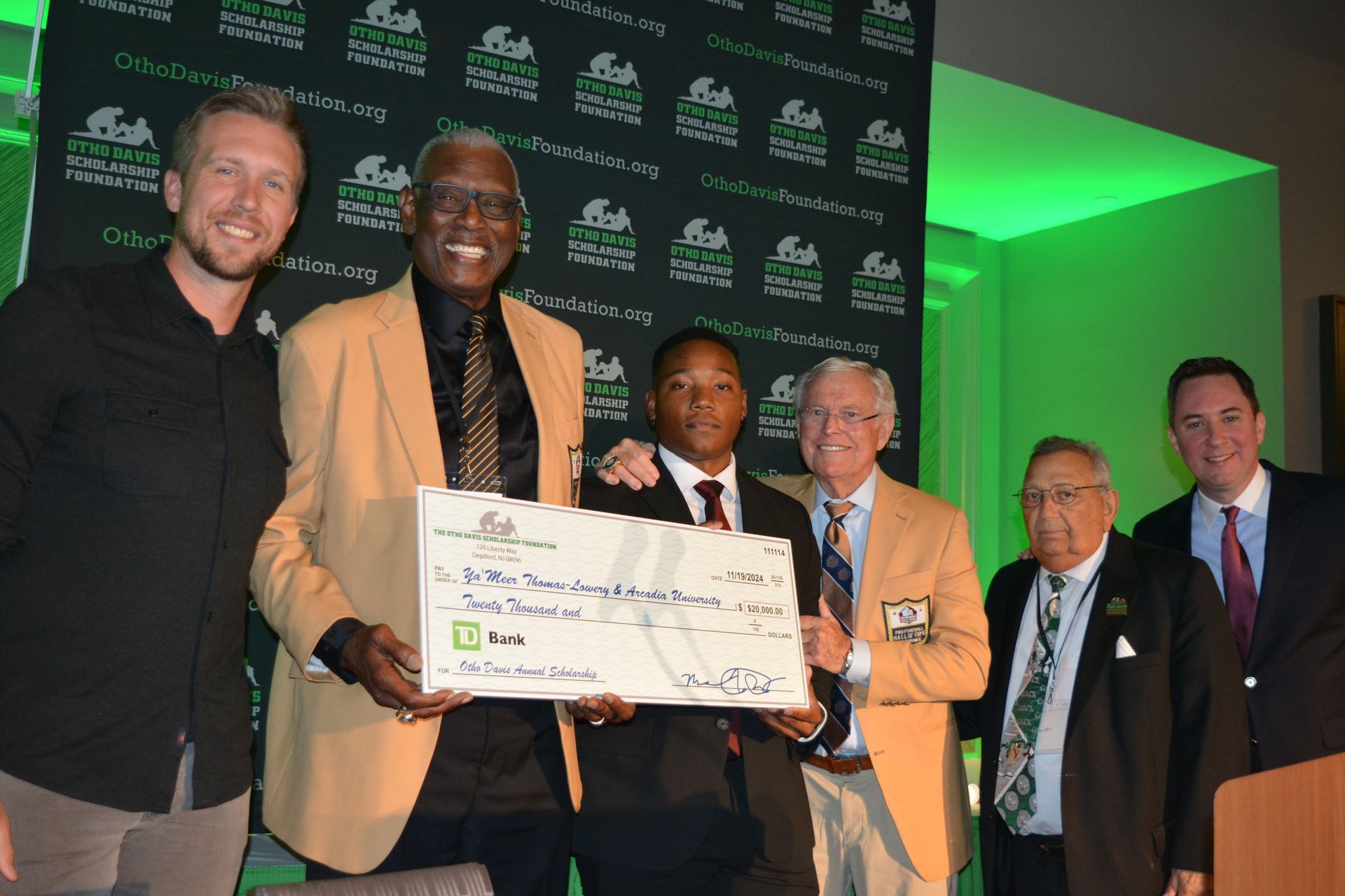 Group of men in jackets presenting a large check to a young person in front of a green backdrop.