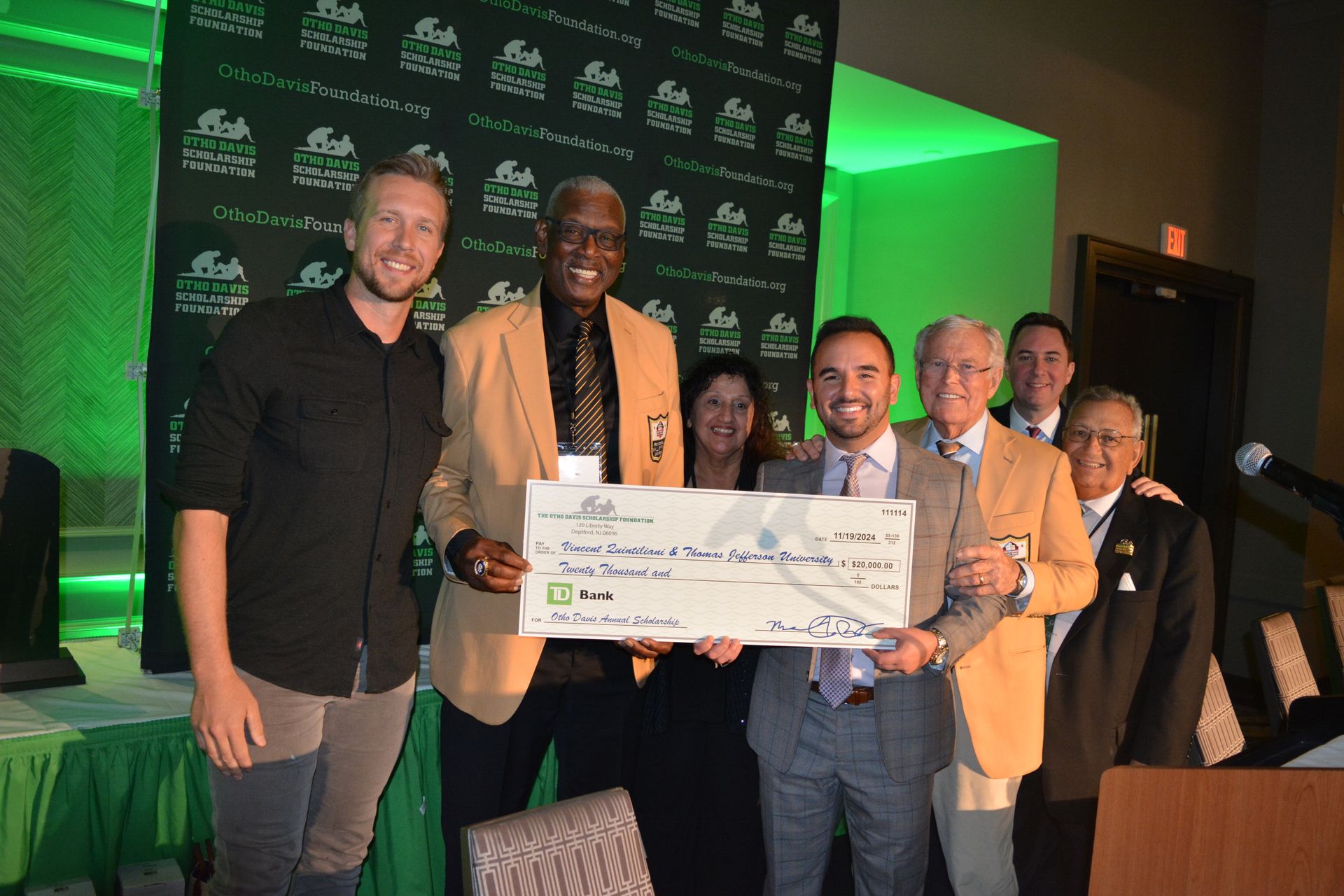 Group of people holding an oversized check, posing in front of a green backdrop at an event.