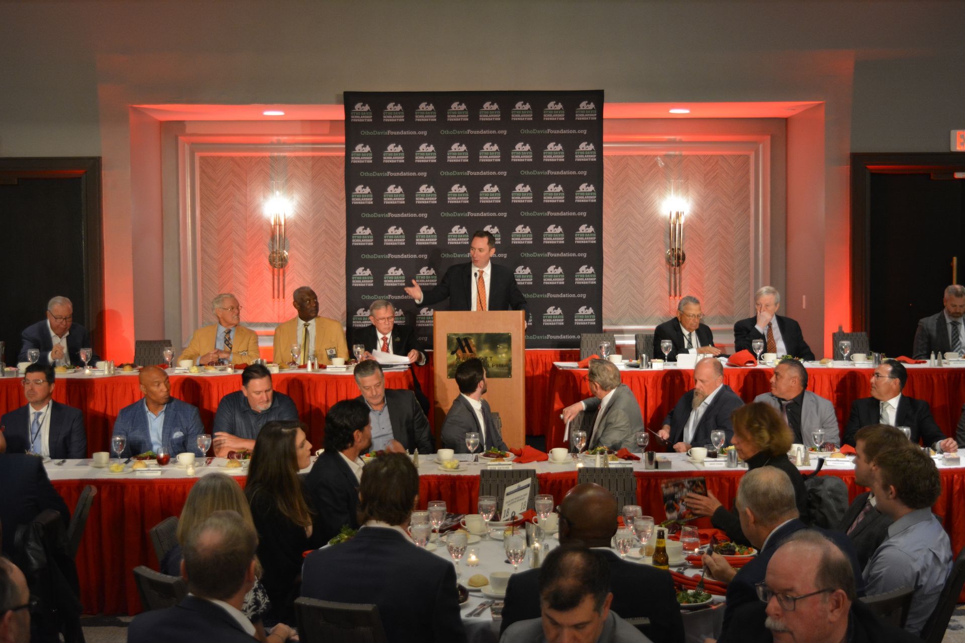 A man is giving a speech in front of a group of people sitting at tables.