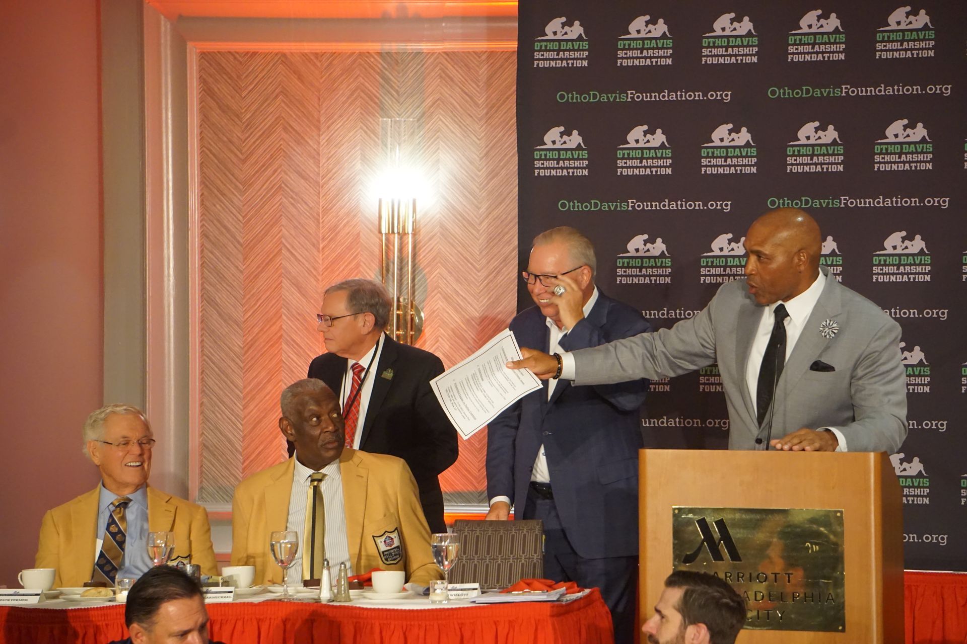 A group of men are sitting at a table with a man standing behind a podium holding a piece of paper.