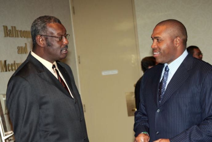 Two men in suits are talking in front of a sign that says 