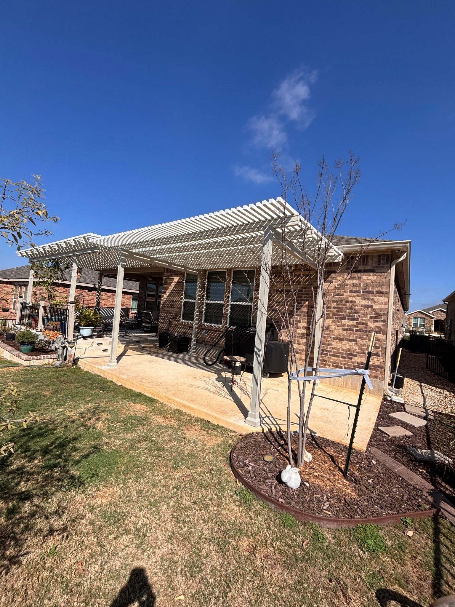 A brown brick house with a large white lattice patio cover over a beige patio, with trees and lawn in the foreground.