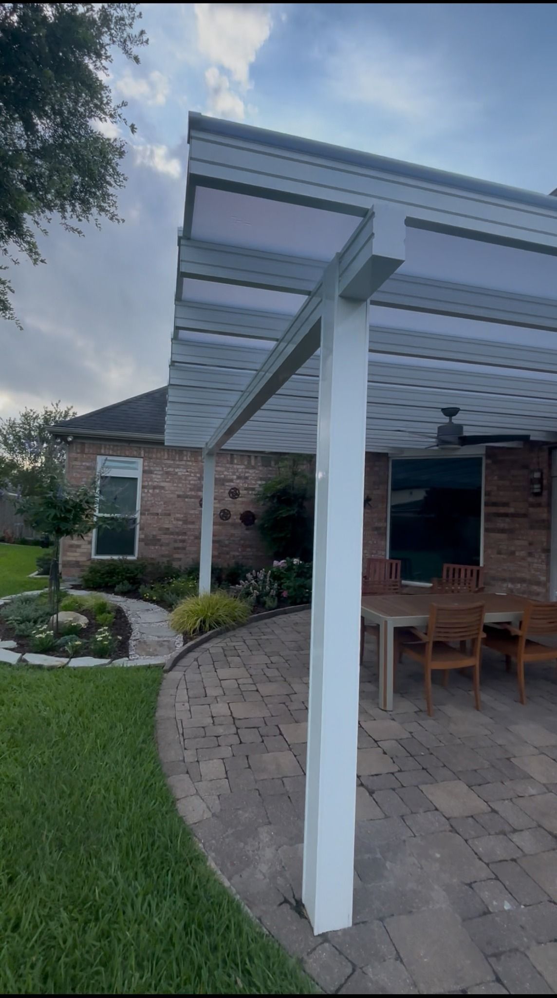 A white pergola shades a brick patio with outdoor furniture next to a brick house and a manicured lawn.