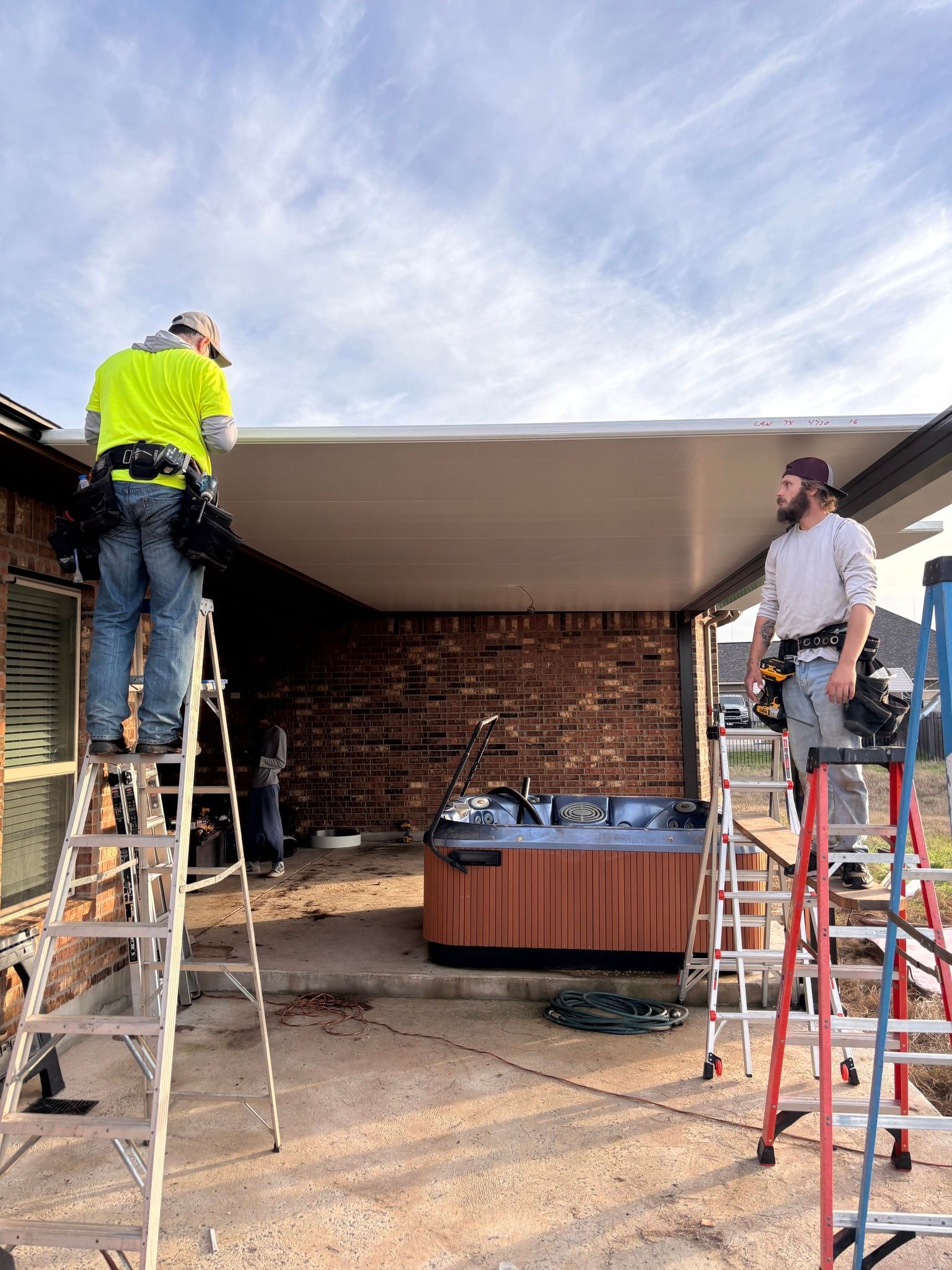 Two construction workers stand on ladders installing a white patio roof cover over a brick patio with a hot tub.