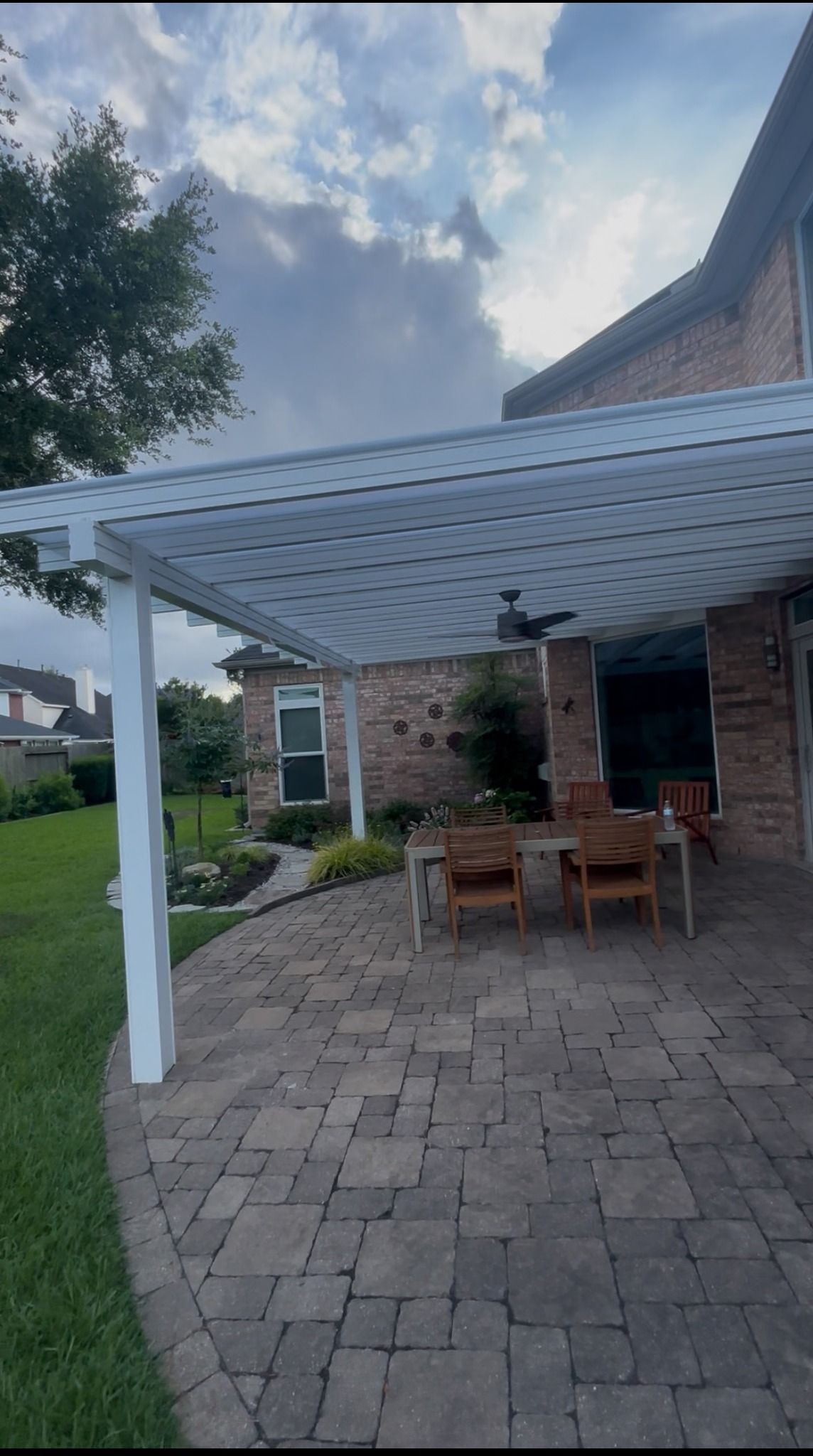 A white patio pergola attached to a brick house covers a dining set on a paver patio overlooking a green backyard.
