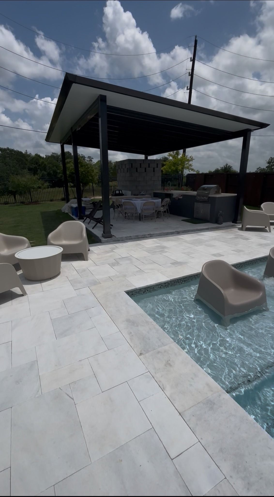 A covered outdoor patio with a dining area next to a swimming pool featuring submerged lounge chairs on a sunny day.