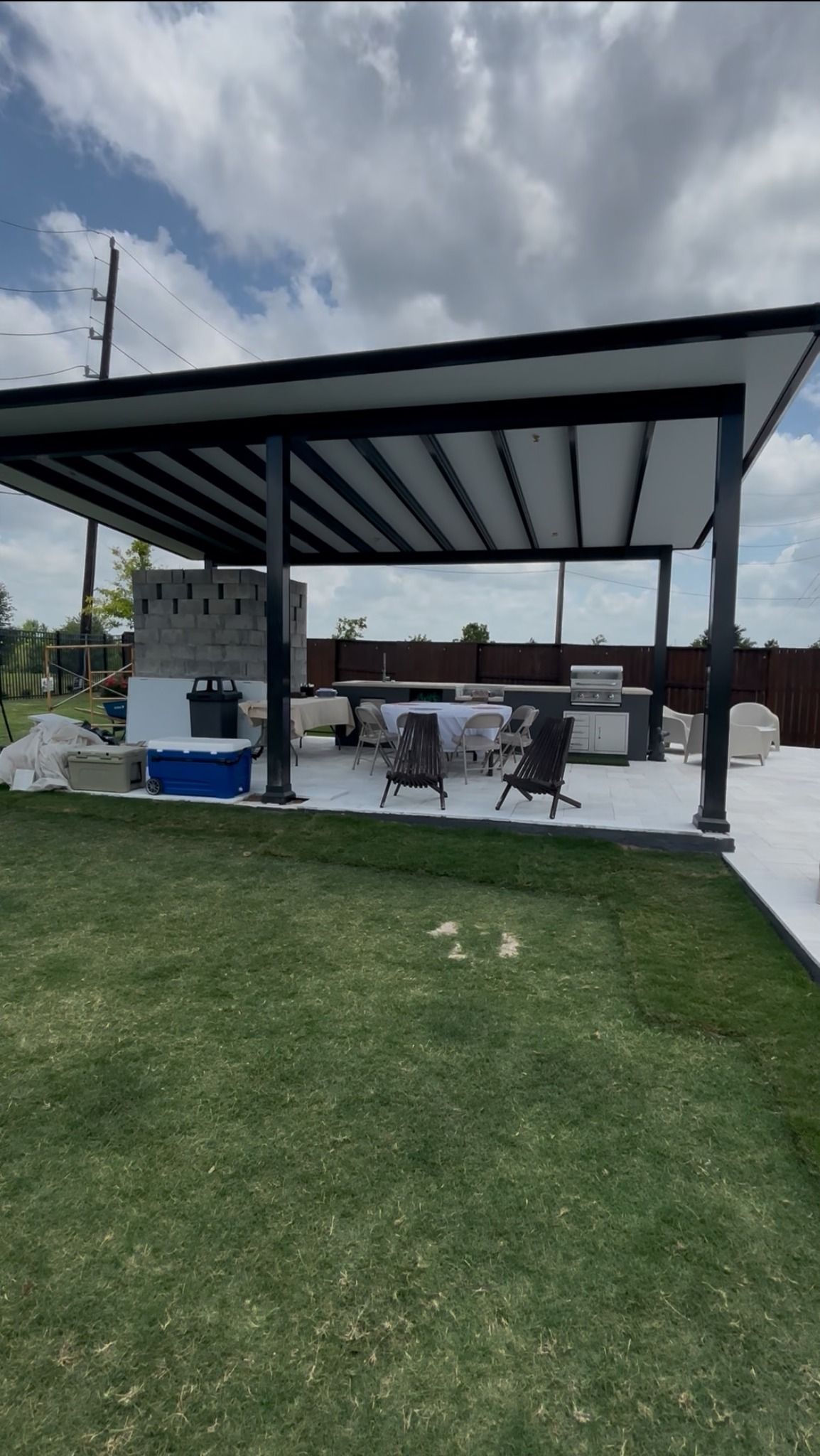 An outdoor patio with a large covered metal roof, an unfinished brick structure, a grill, and seating on a sunny day.