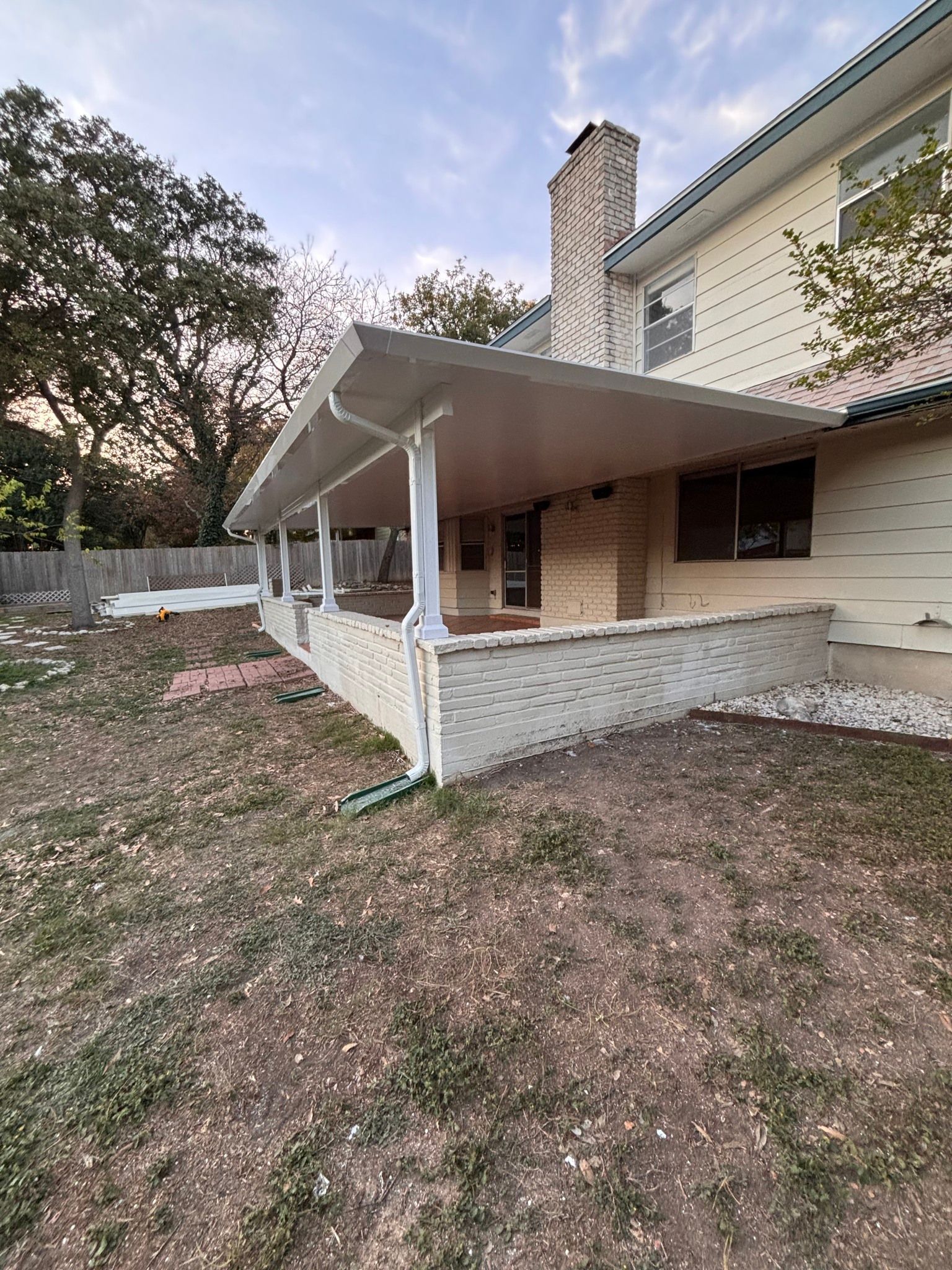 A patio with a white covered roof and brick half-wall attached to the side of a two-story beige house.