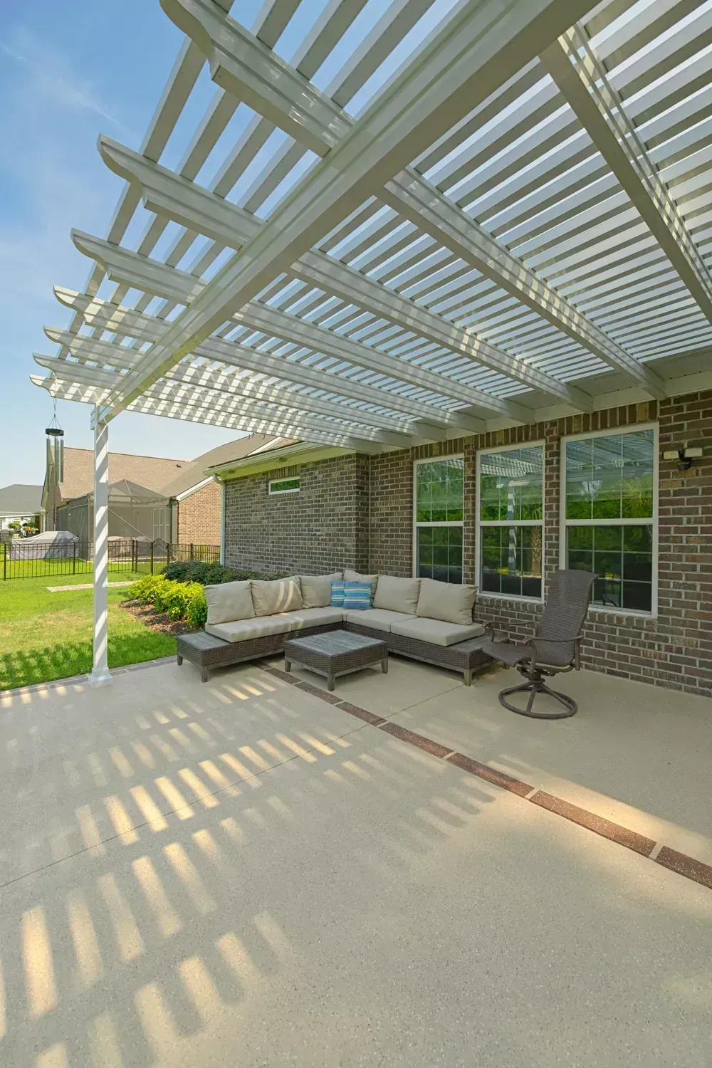 A patio with a white pergola covering a sectional sofa, coffee table, and chair against a brick house exterior.