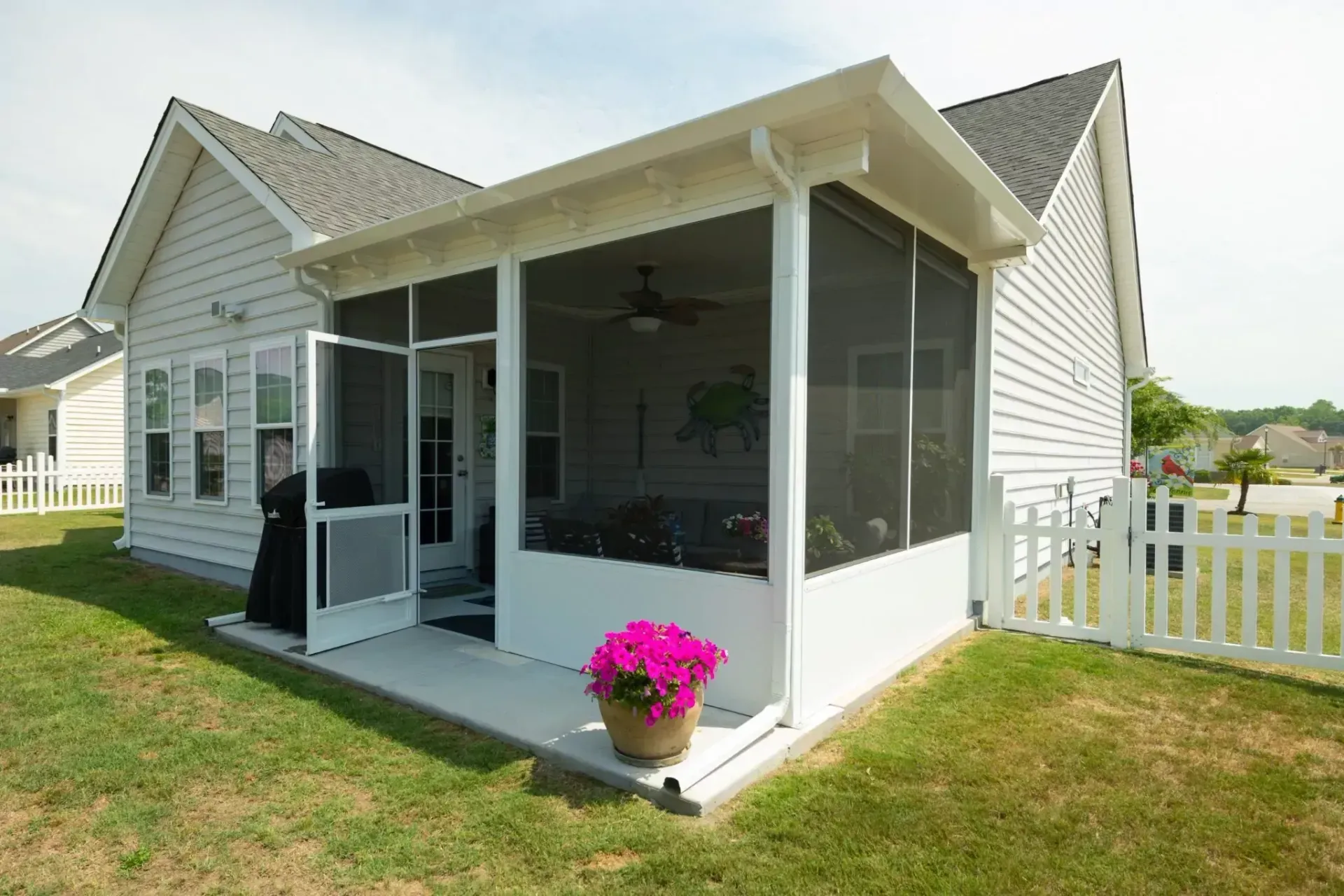 A light-colored house with a screened-in porch, a potted pink flower, and a white picket fence on a sunny day.
