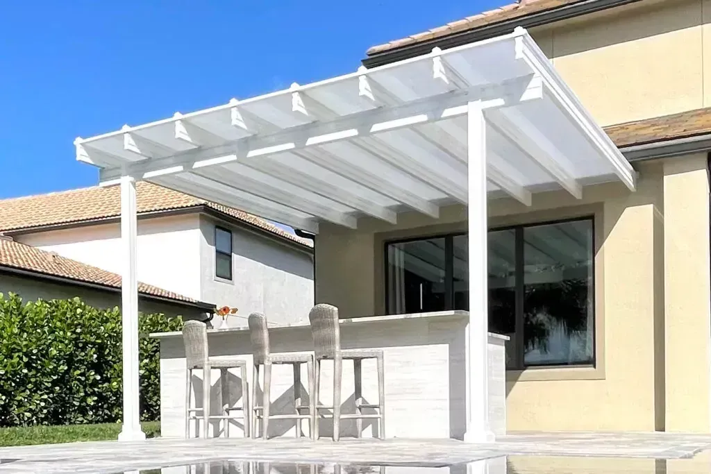 A white pergola with a translucent roof covers an outdoor stone bar with three tall stools on a sunny patio.