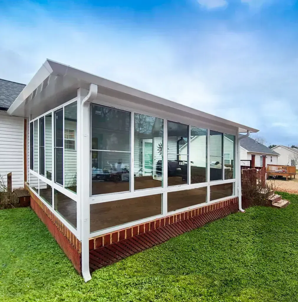 A white, glass-enclosed sunroom addition with a brick base attached to the side of a house, set in a grassy yard.