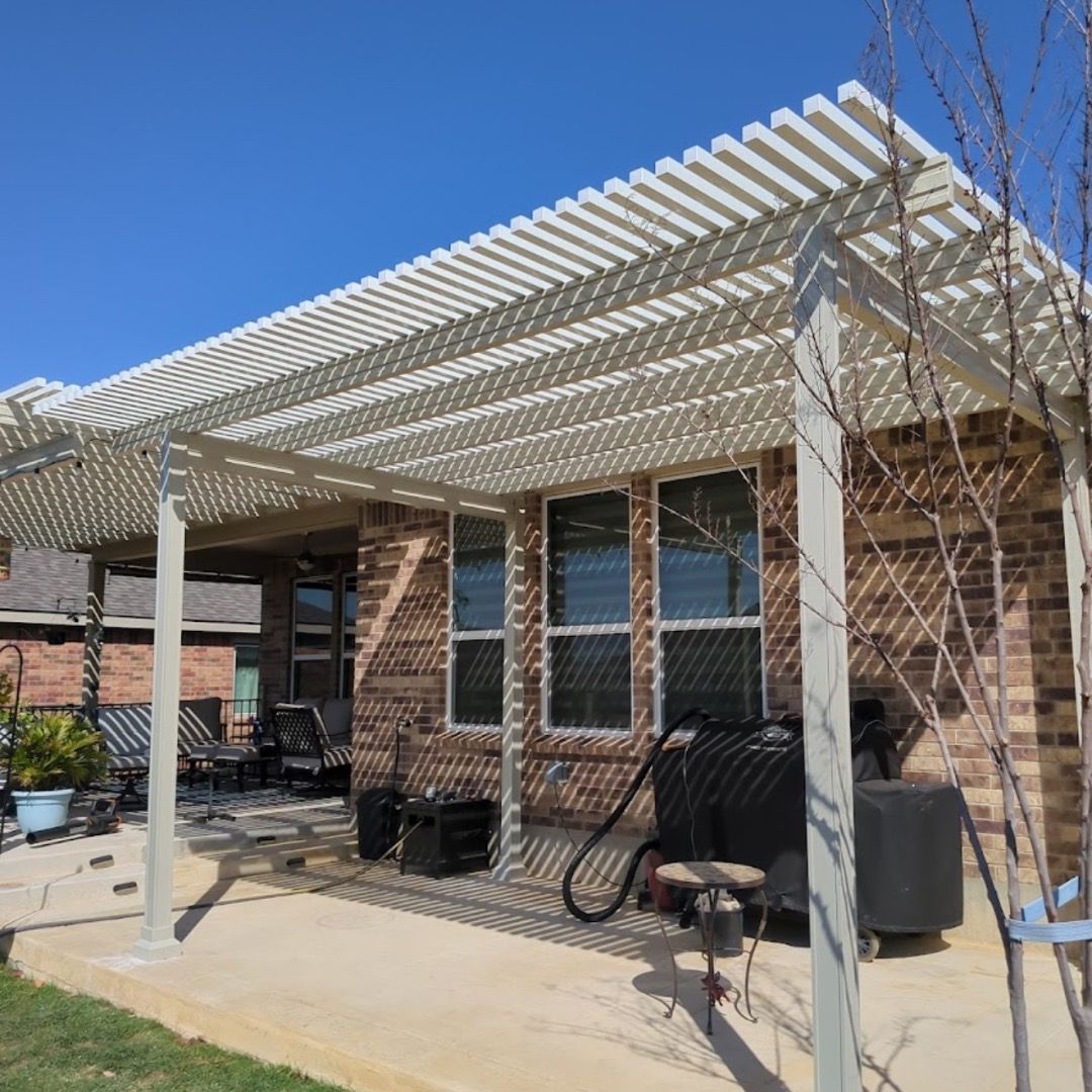 A white pergola attached to a brick house covers a patio with outdoor furniture, a grill, and a small sapling nearby.