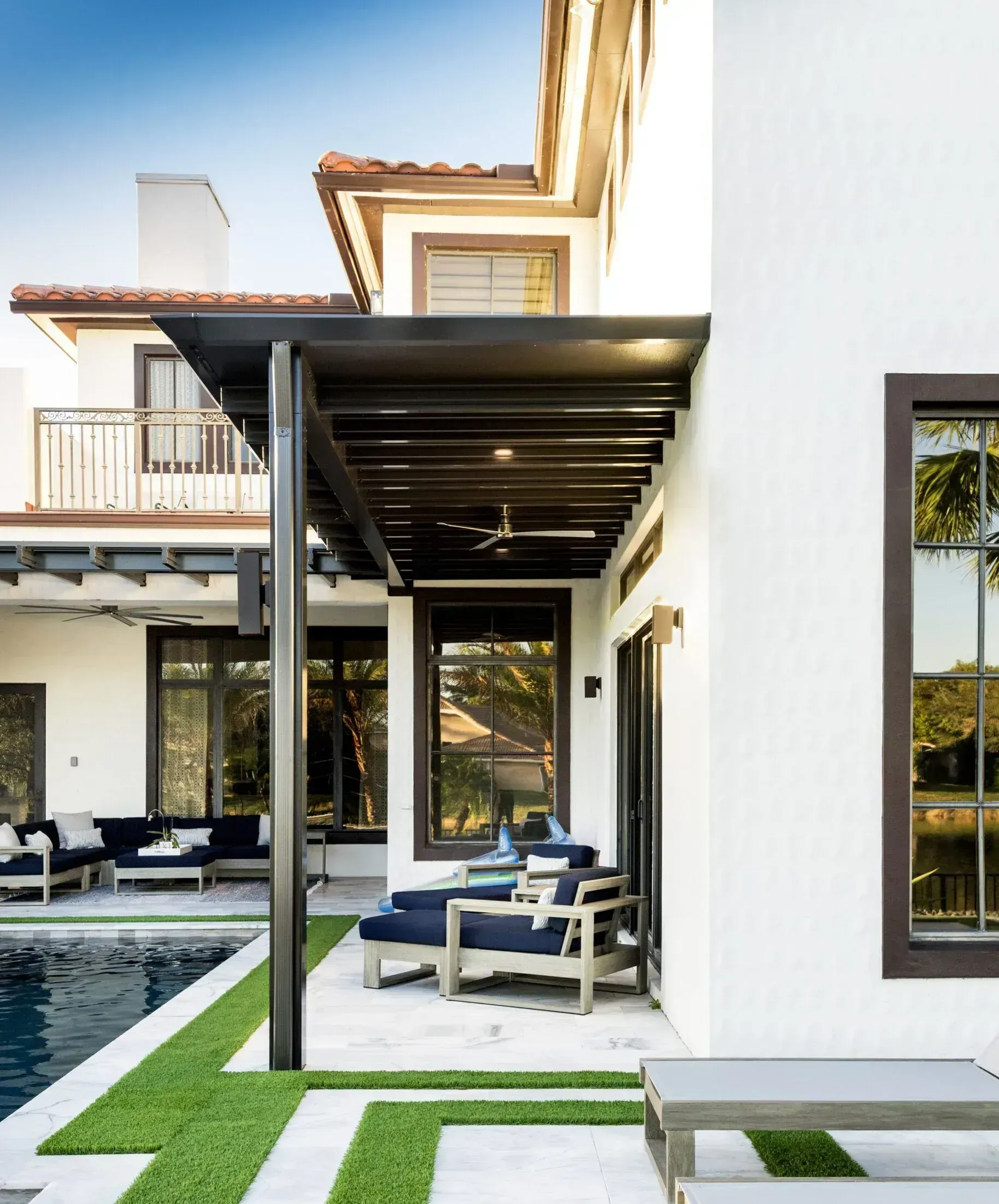 A white house with dark framed windows and a covered patio overlooking a swimming pool with lounge chairs on stone pavers.