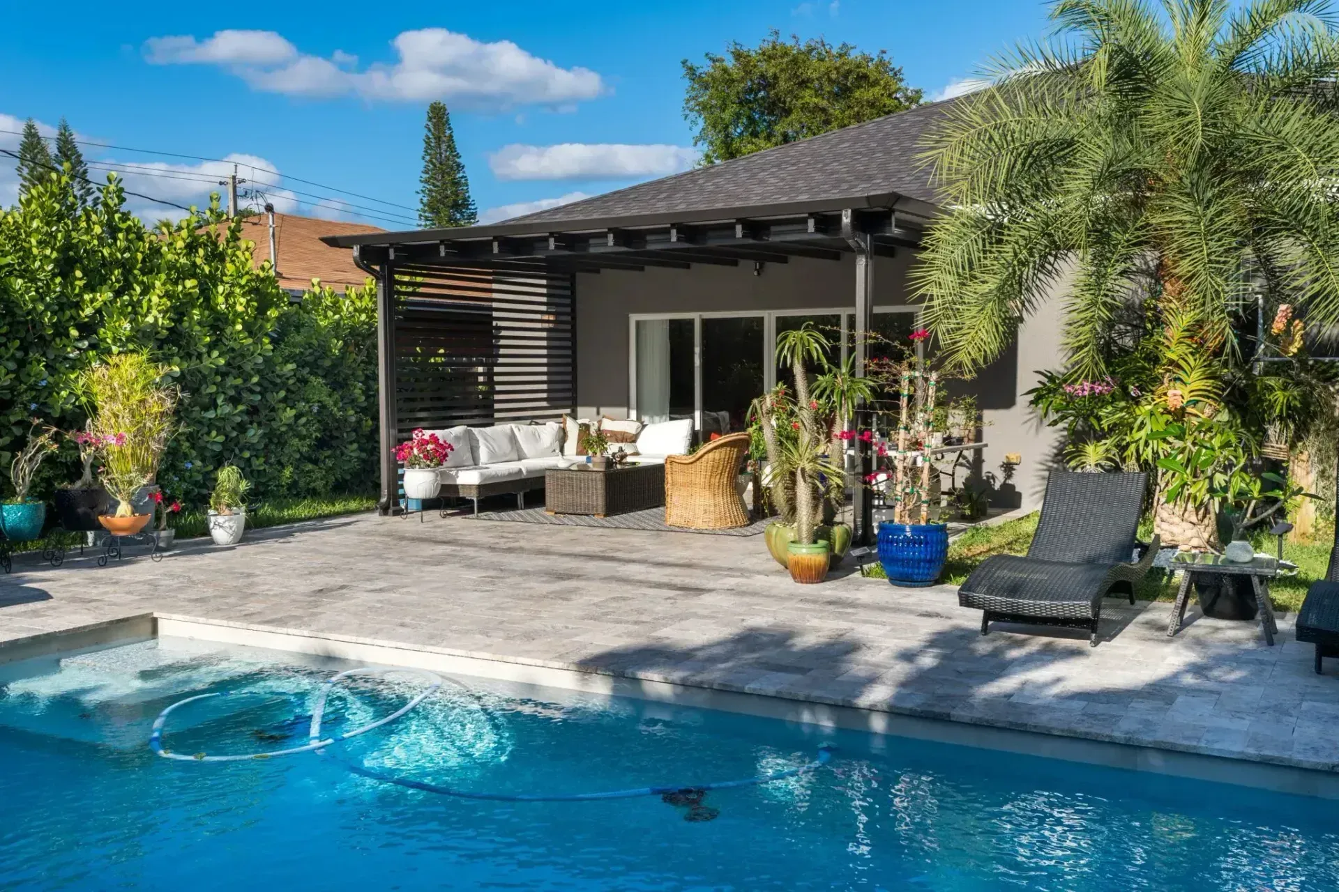 A bright swimming pool patio area featuring outdoor seating, a shaded pergola, potted plants, and a lounge chair.