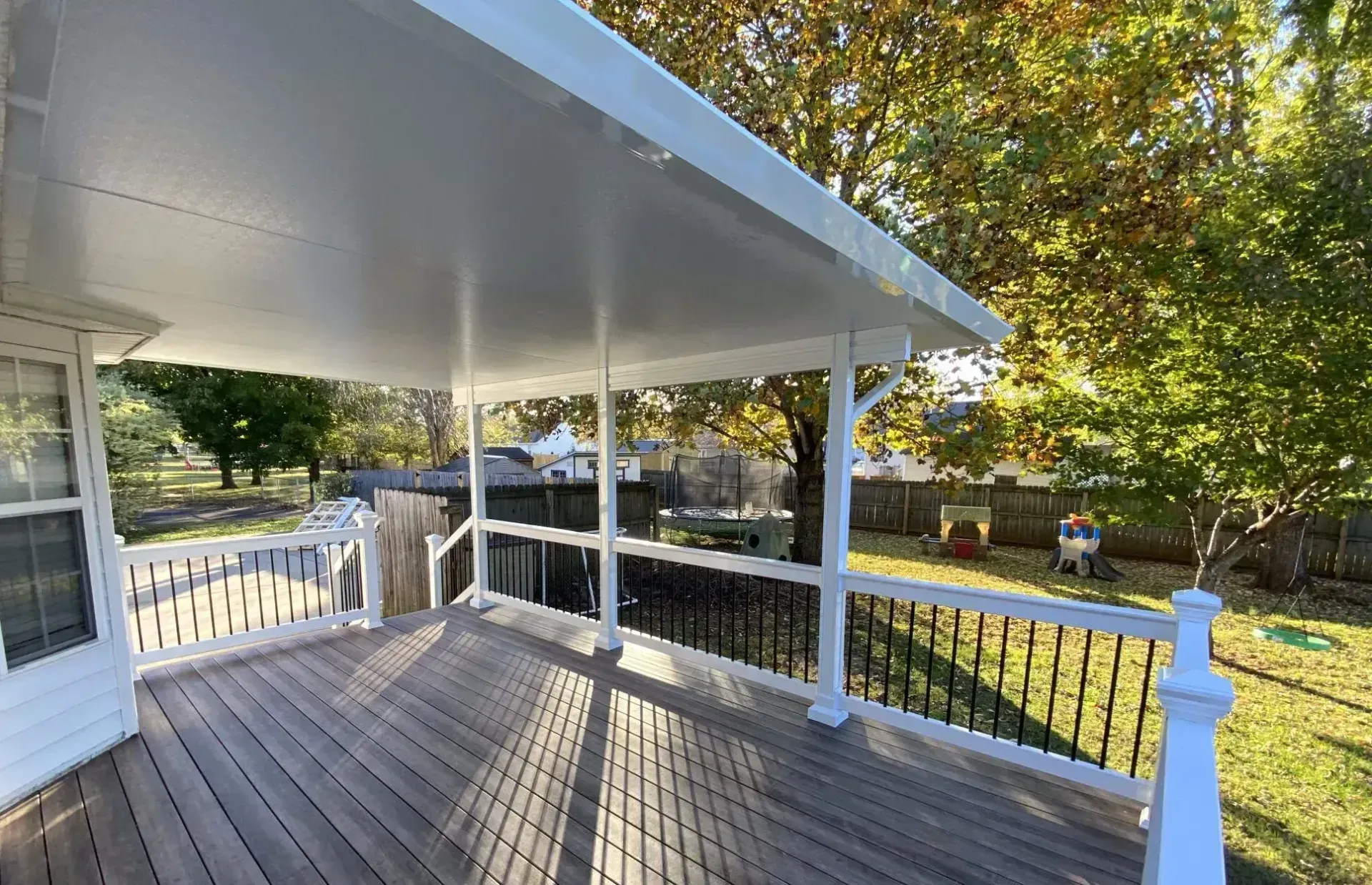 A covered wooden deck with white railings and support posts, overlooking a backyard with trees and lawn.