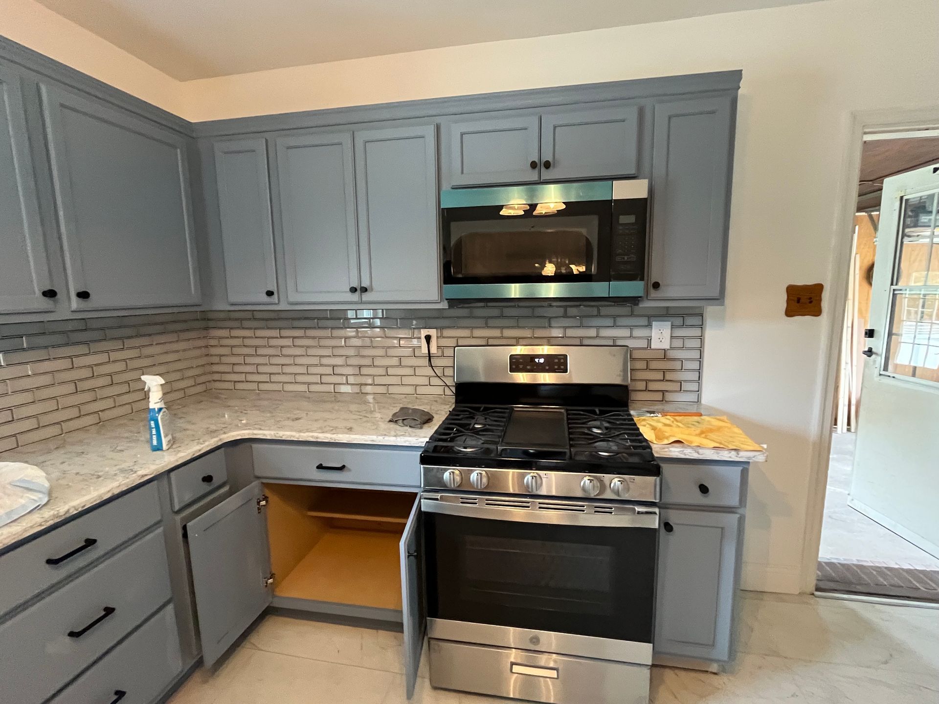 Gray kitchen with gas stove, microwave, and blue cabinets, plus open under-sink cabinet.