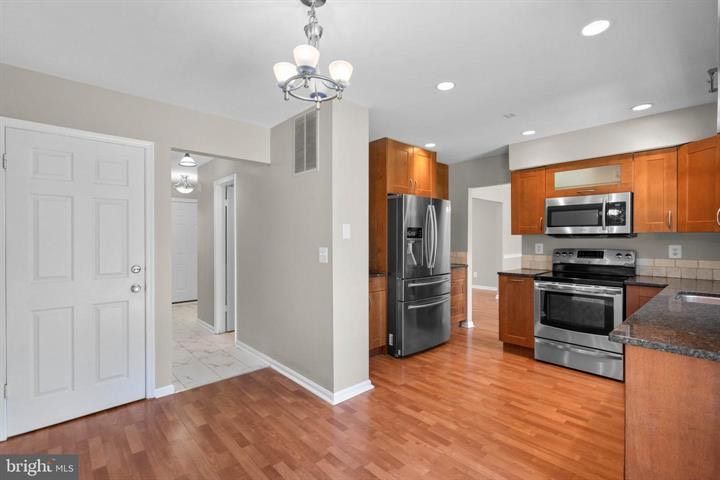 Bright kitchen with stainless steel appliances, wood cabinets, hardwood floors, and an adjacent hallway.