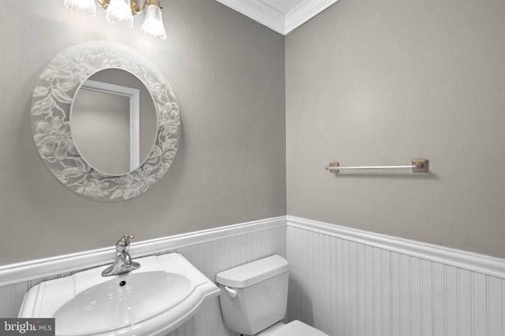 Small bathroom with pedestal sink, toilet, round mirror, and wainscoting in gray and white.