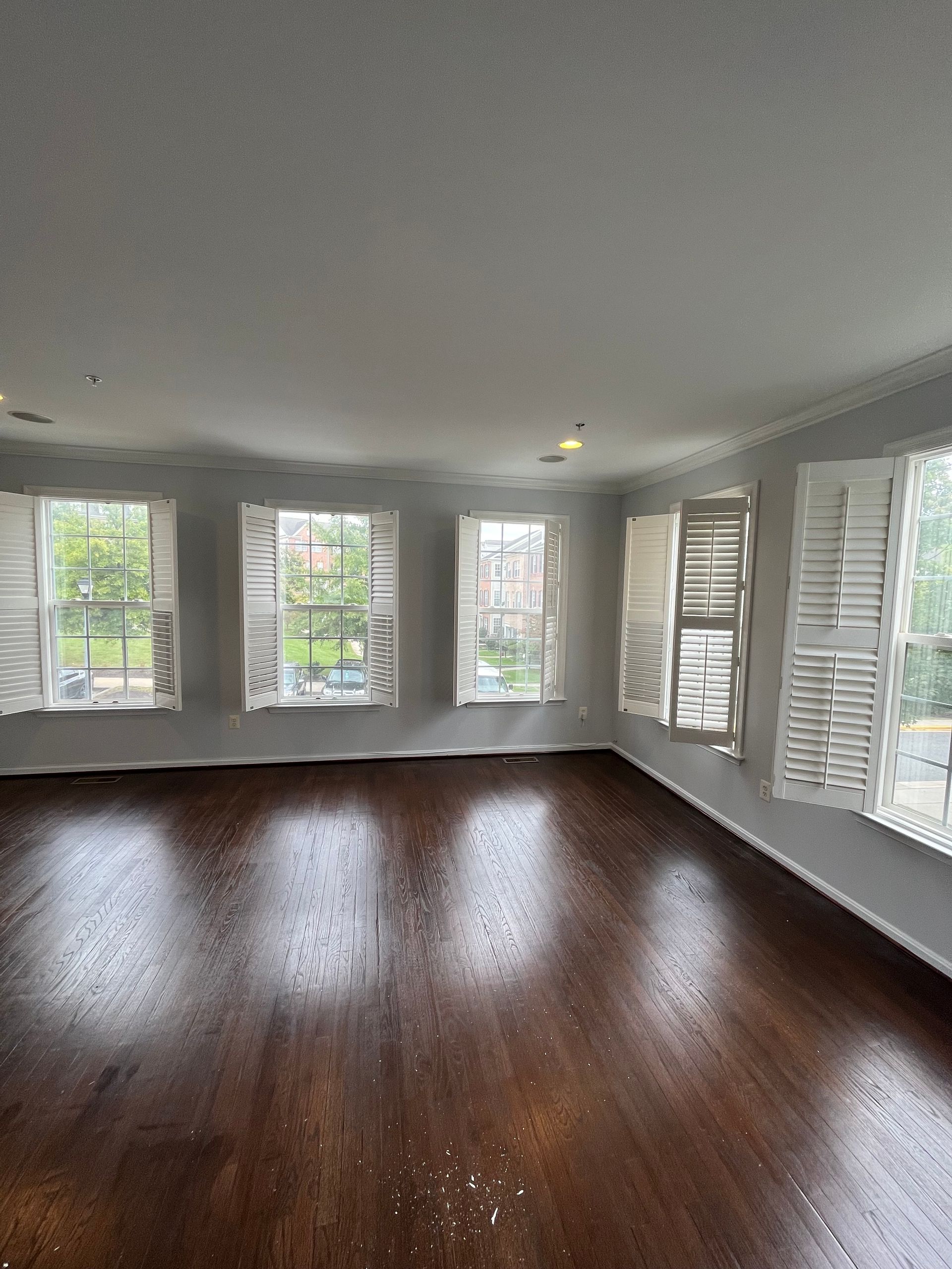 Empty room with dark hardwood floors, white walls, and large windows with shutters.