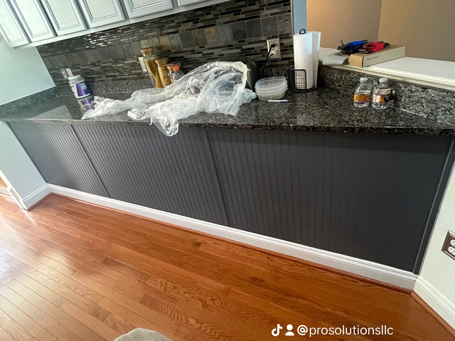 Kitchen counter with black granite top, dark paneled cabinets, and hardwood floor, with items and plastic wrap on the counter