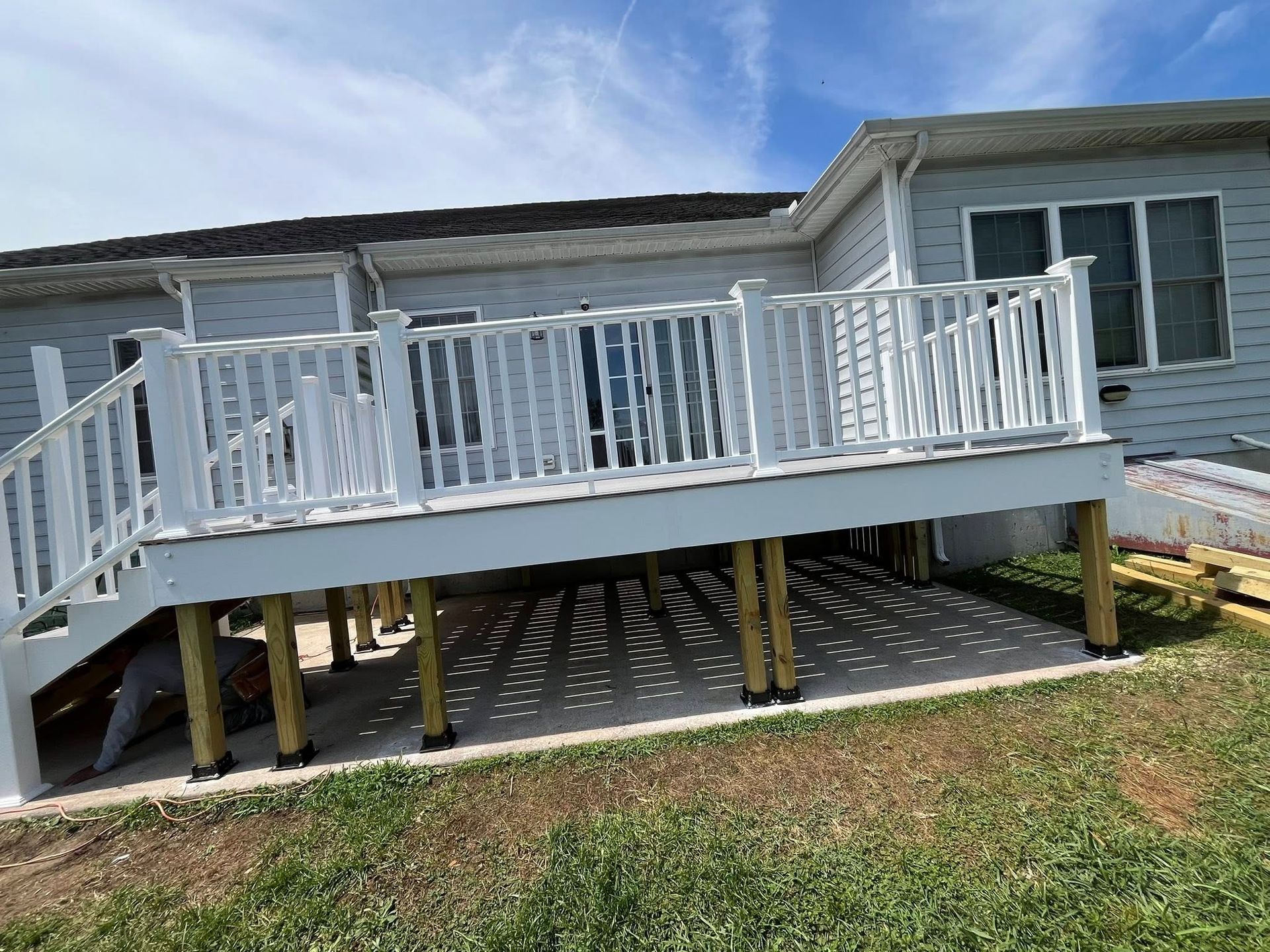 White deck with white railing attached to a light-colored house. Wooden posts support the deck over concrete.