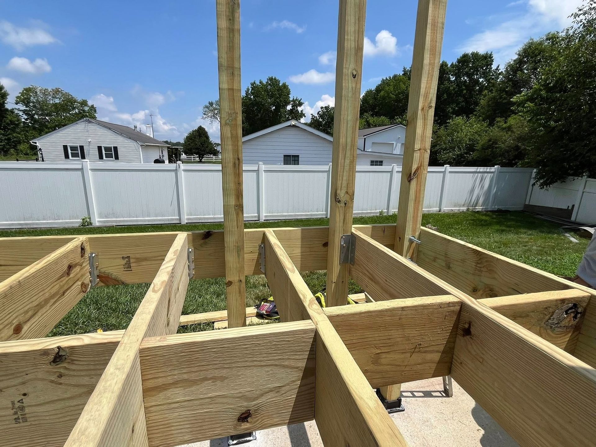 Framed wooden deck under construction; vertical support posts rise. White fence, houses, green grass visible.