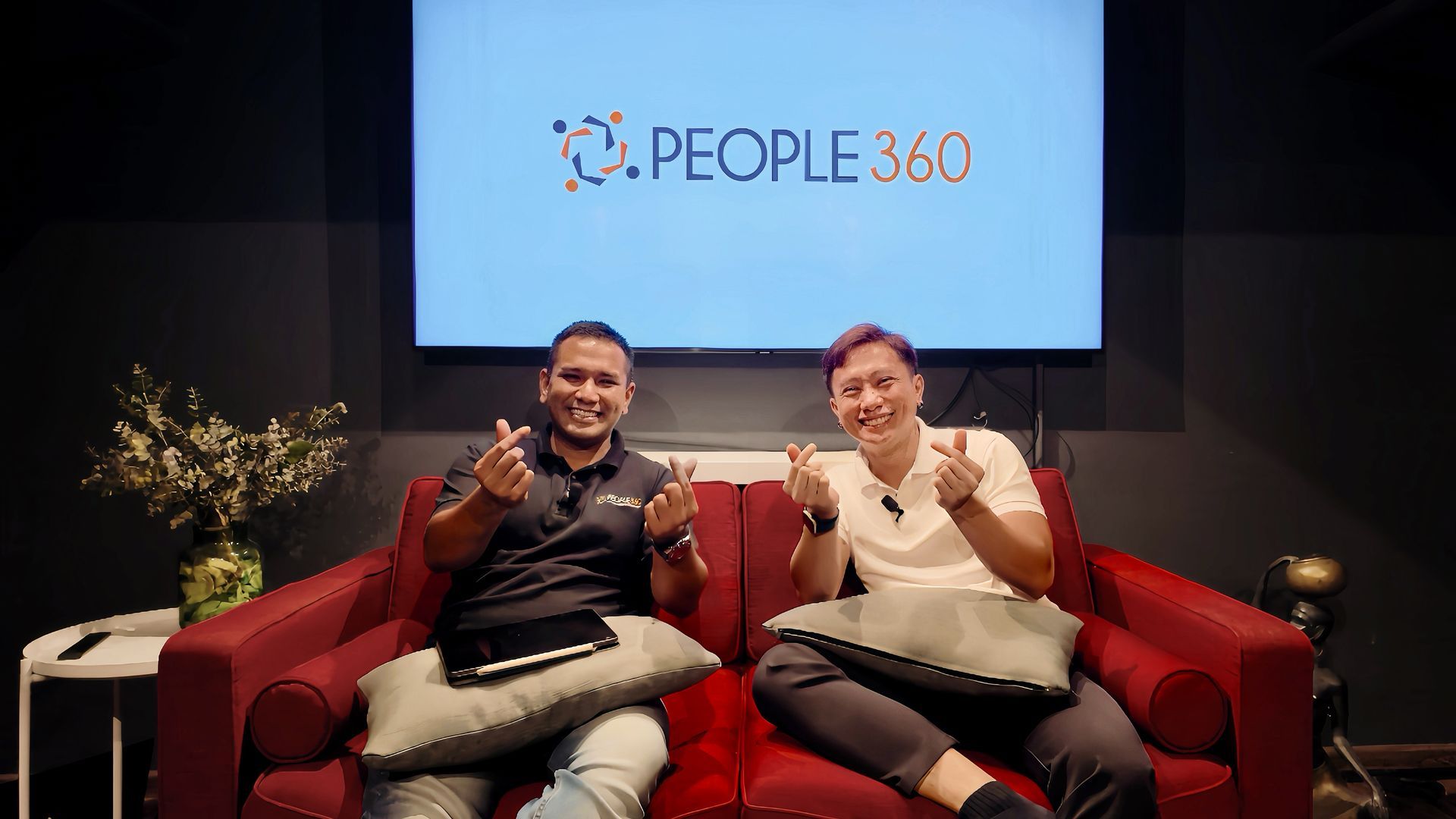 Two men are sitting on a red couch giving thumbs up.