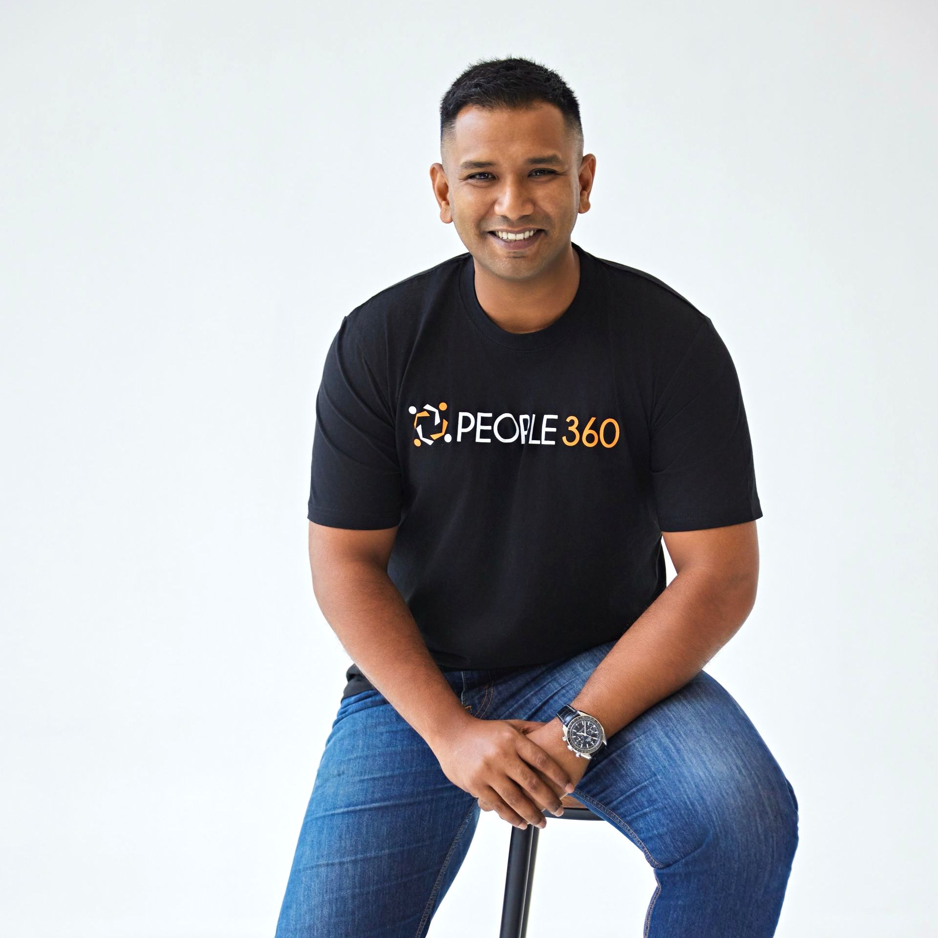 A man is sitting on a stool wearing a pecile 360 shirt