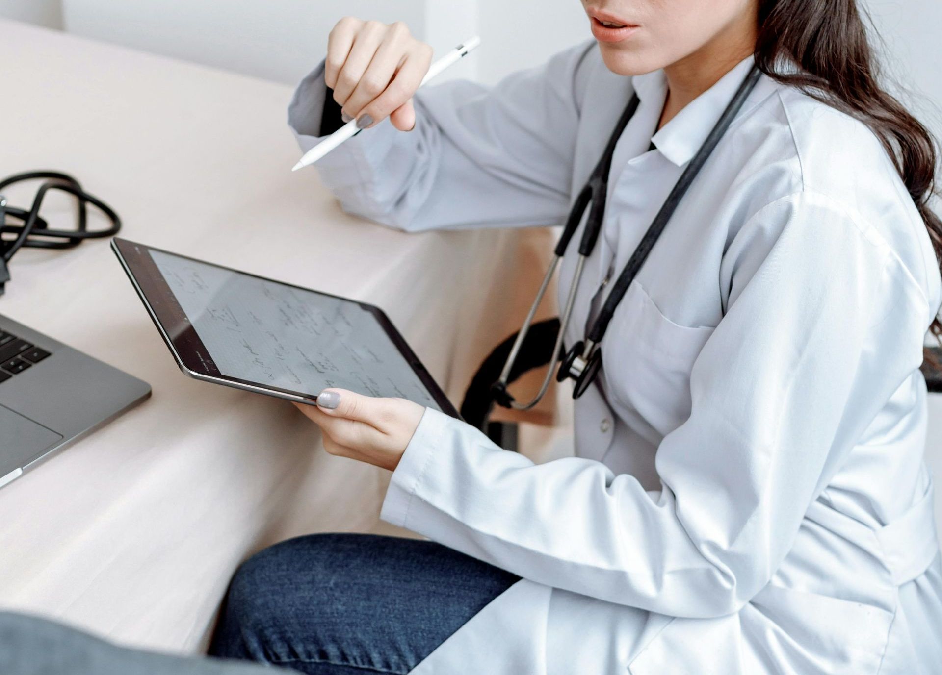 A female doctor is sitting at a desk holding a tablet and a pen.