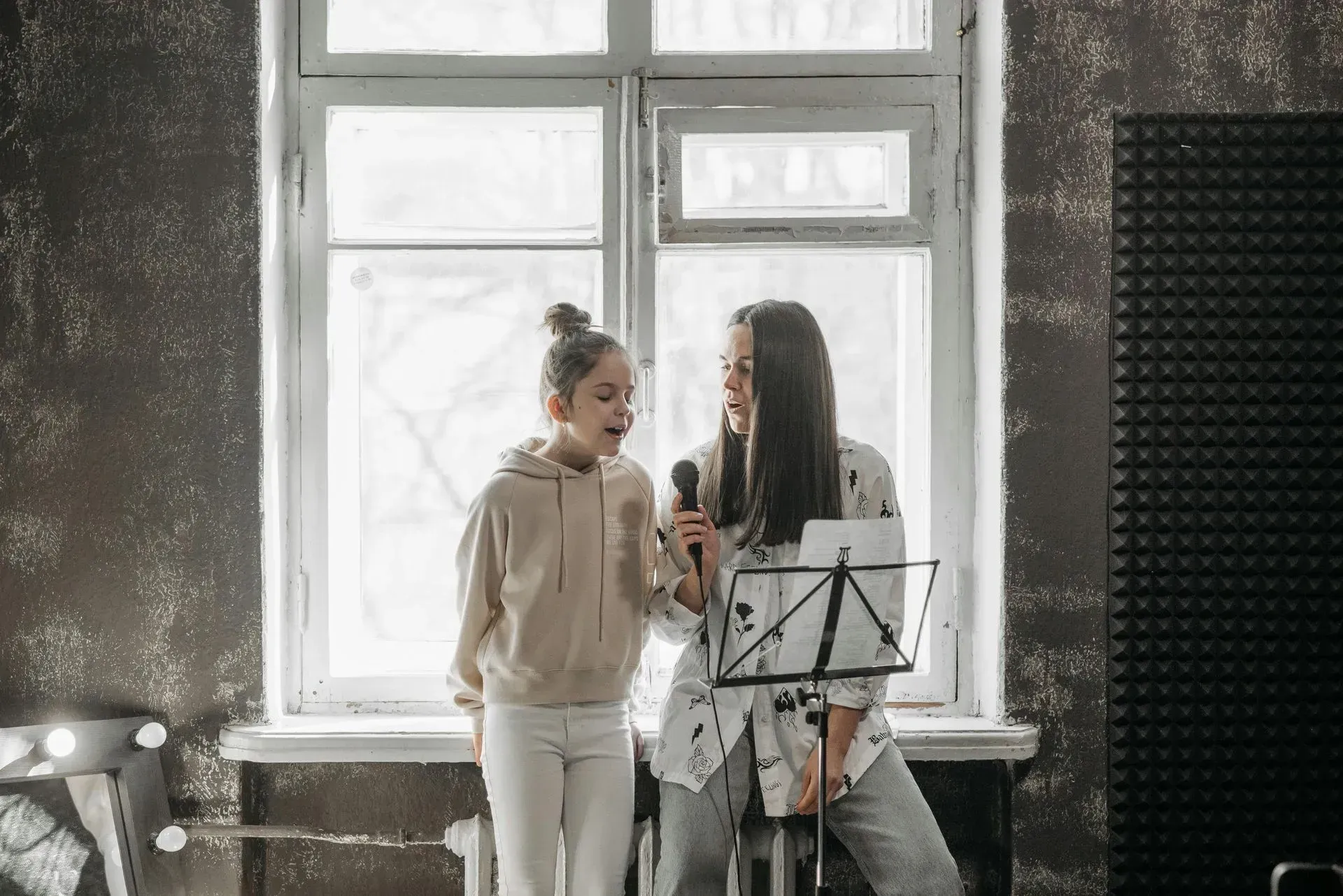 Two women are singing into microphones in front of a window.