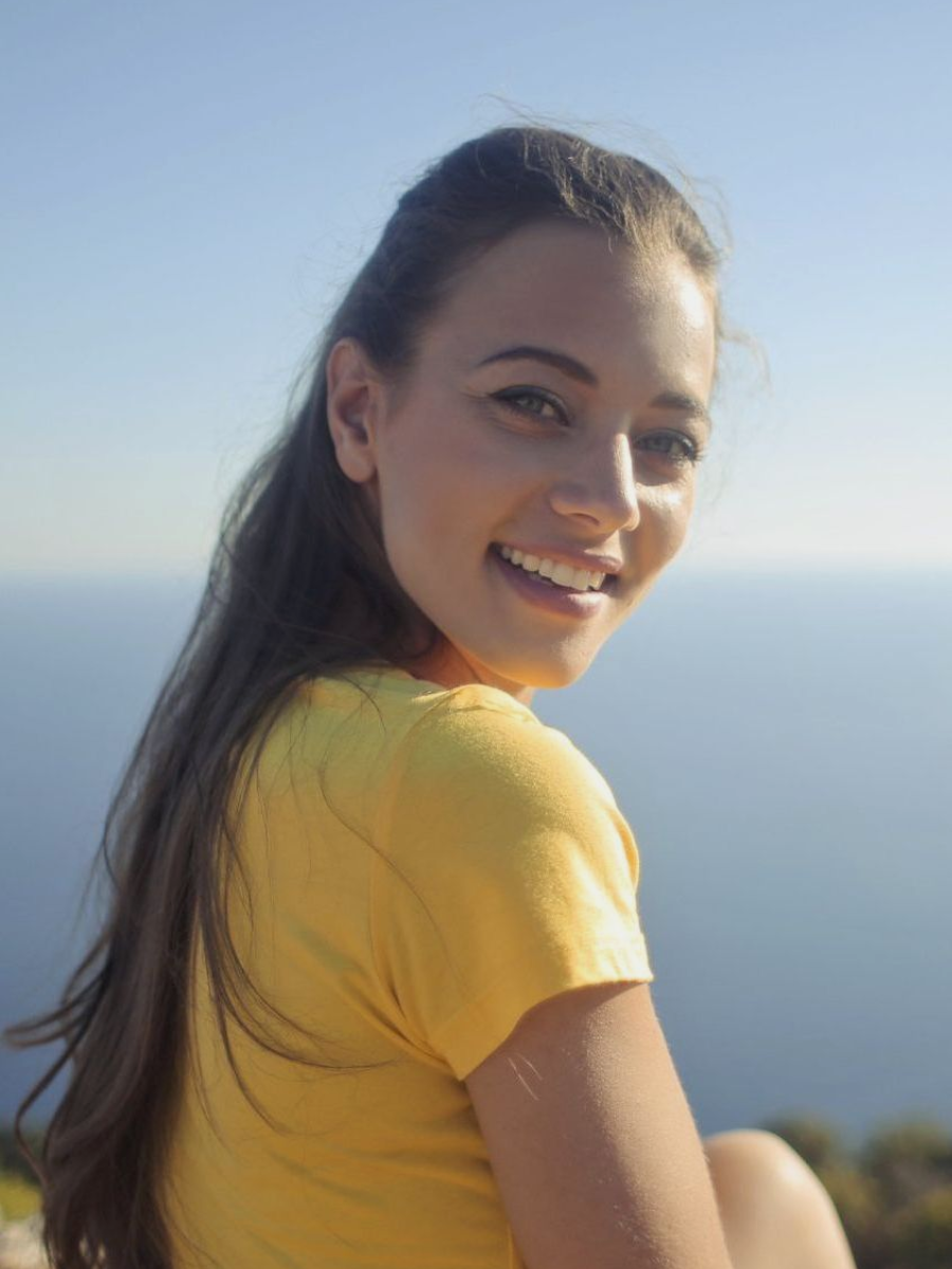Woman with brown hair smiles, looking over her shoulder. Wears a yellow shirt, with a sea view backdrop.