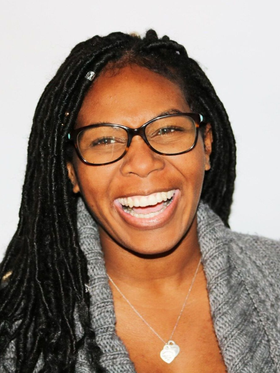 Woman with locs, glasses, and grey sweater, smiling widely.