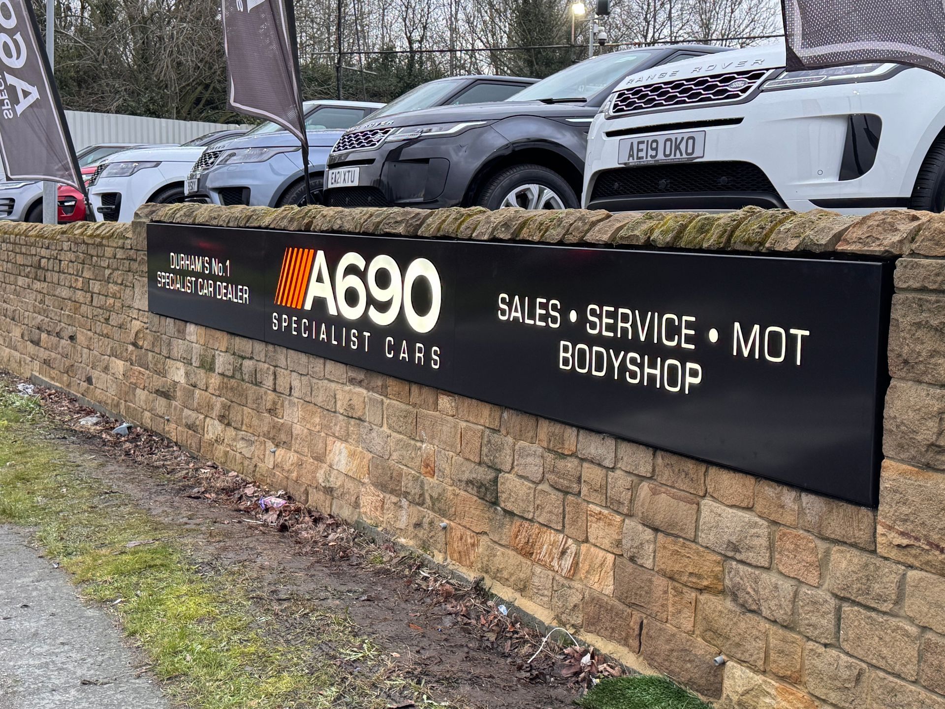 Sign for A690 First Cars, displaying services: sales, service, MOT, and bodyshop, on a stone wall.