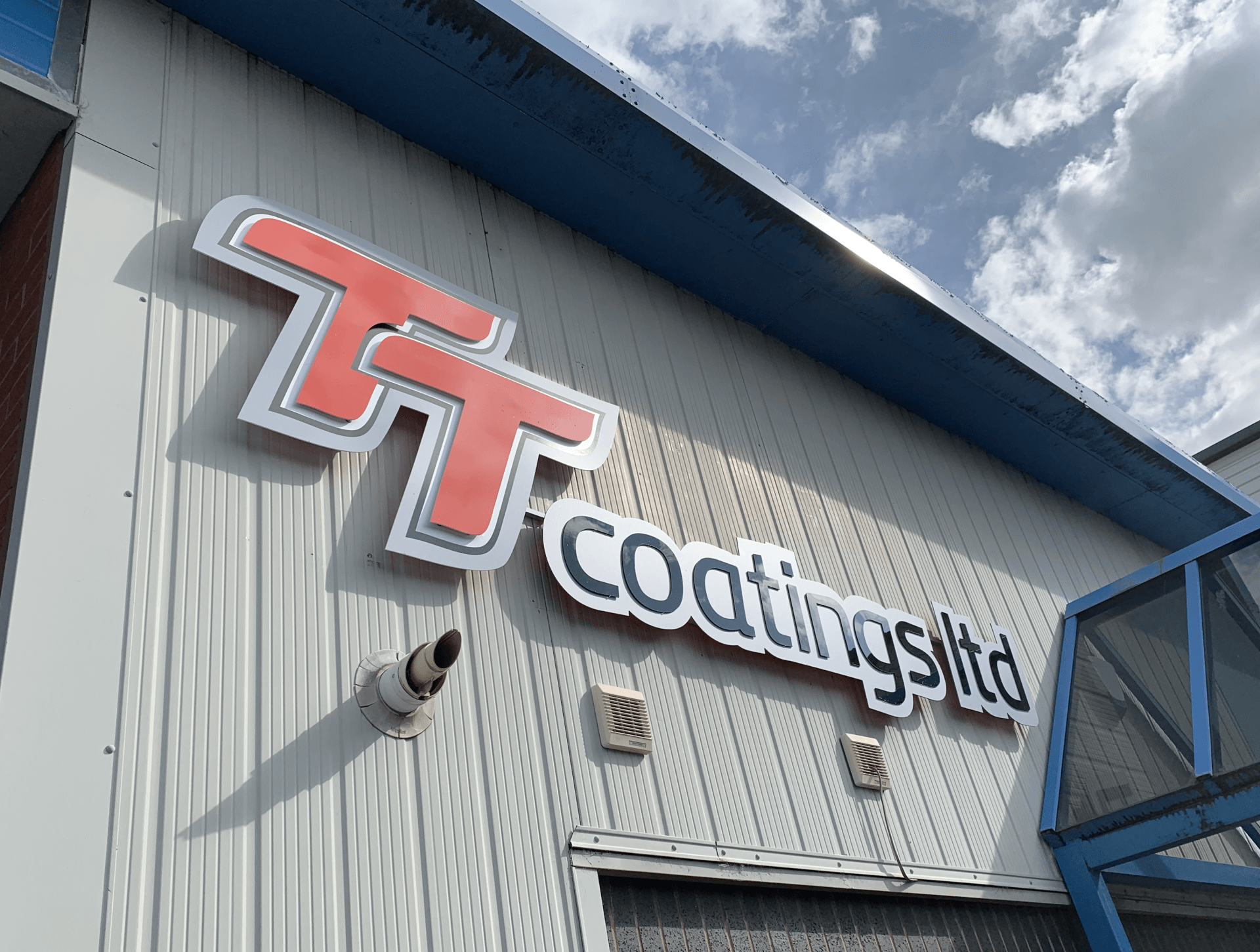 TT Coatings Ltd sign on a white industrial building. Red and silver logo. Blue trim.