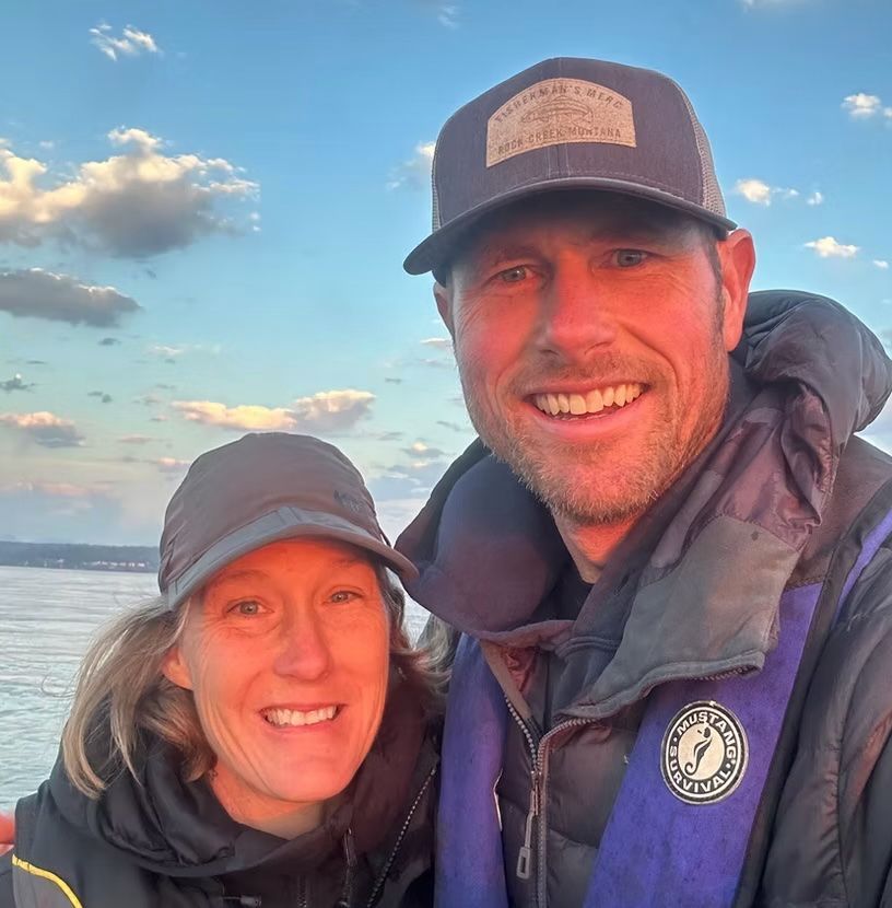 Couple smiling, bundled up, wearing hats and life vests, posing outdoors near water at sunset.