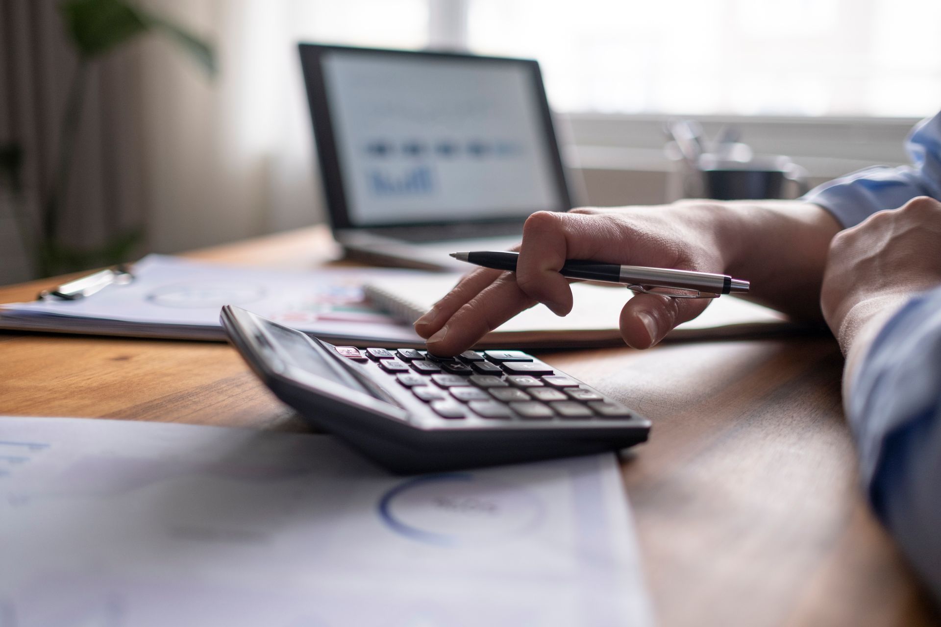 Hands using a calculator, with laptop and documents on a desk, presumably doing financial calculations.