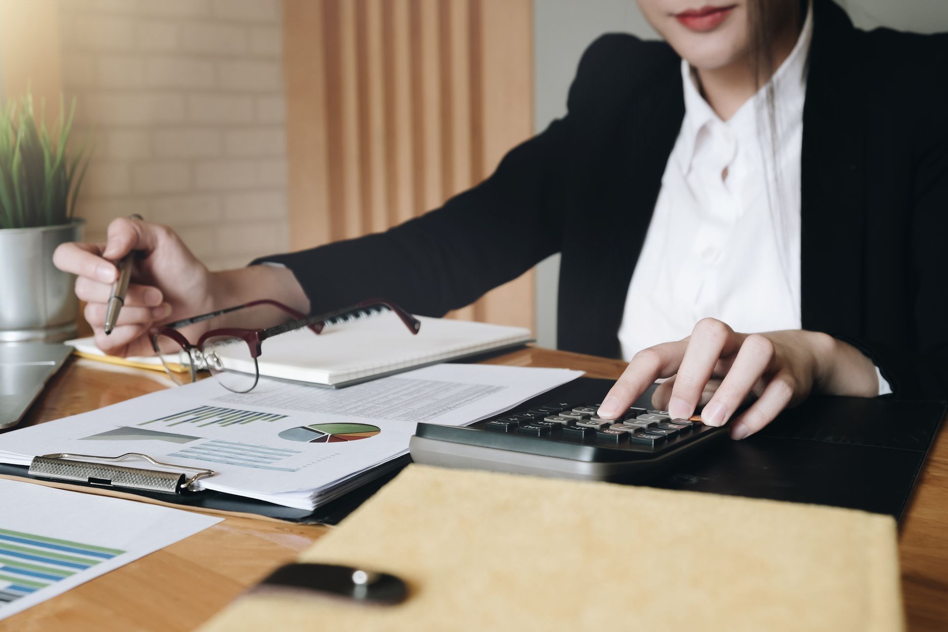 Woman in a suit uses a calculator at a desk, reviewing financial documents and holding glasses.
