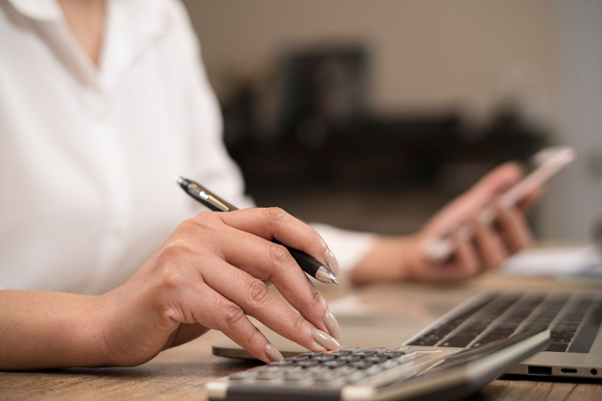 Woman using a laptop with a pen in hand, also holding a phone, in an office setting.