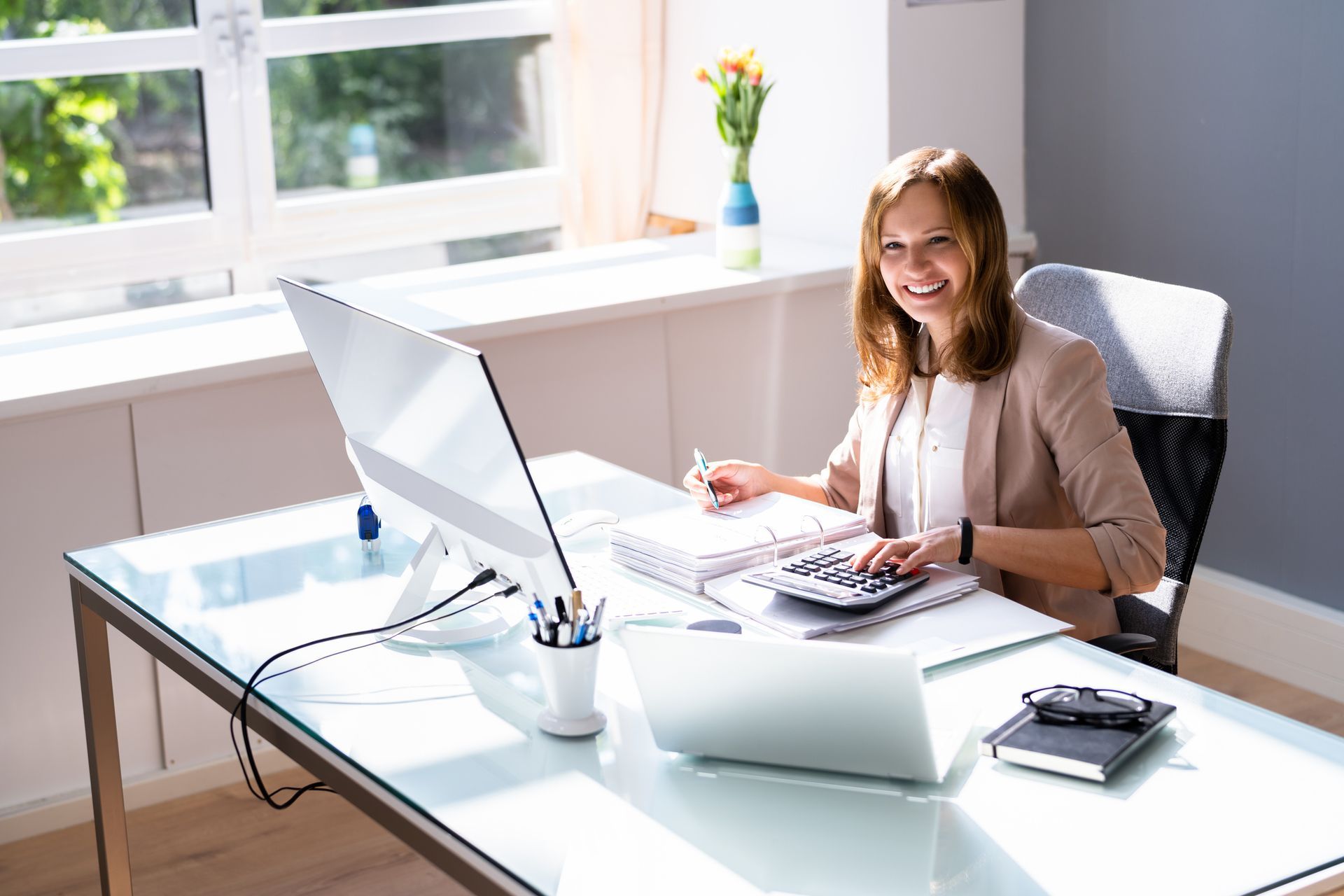 Woman smiles while working at a glass desk with a computer, calculator, and notebook in an office.