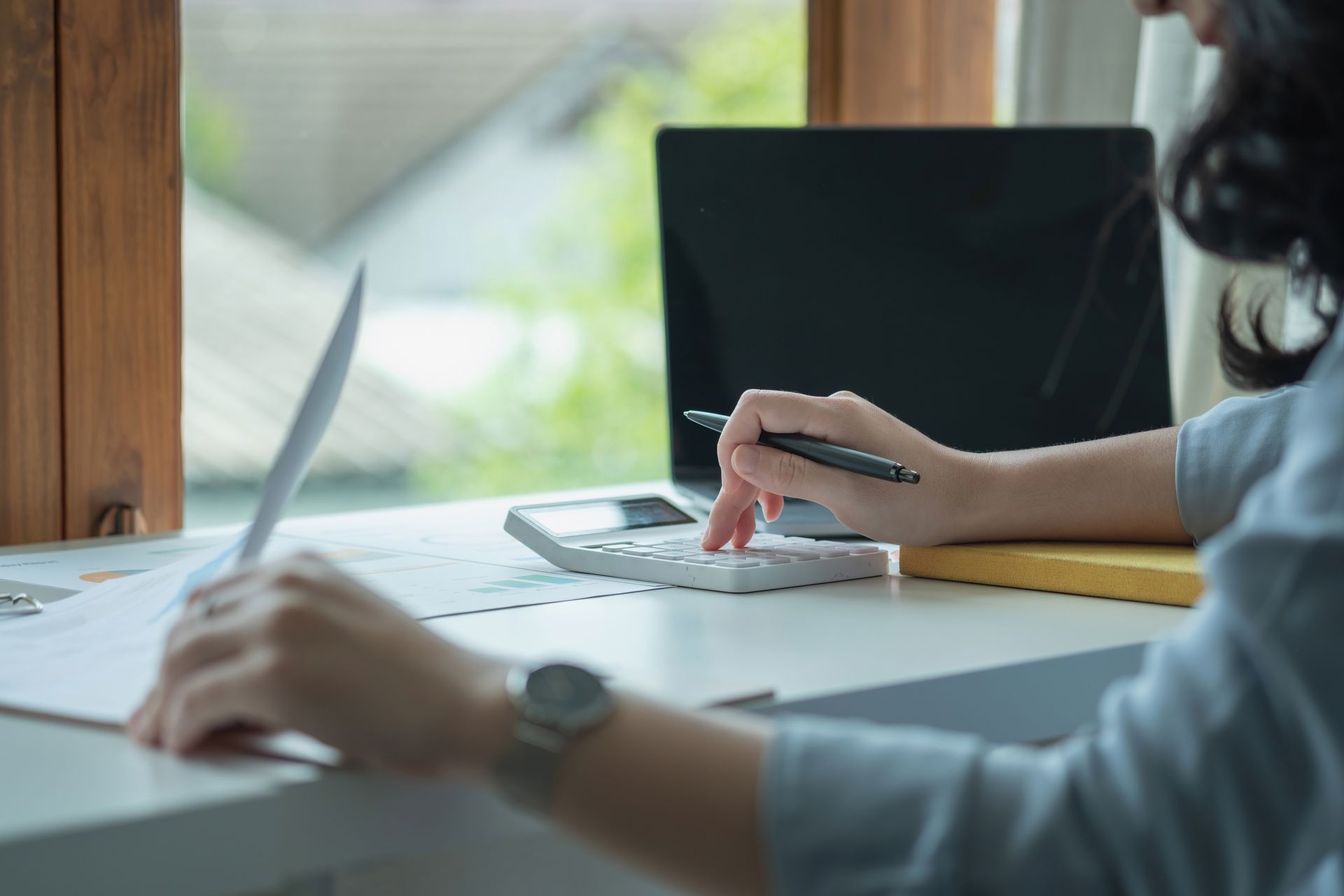 Woman using calculator and reviewing papers at a desk with laptop and window.