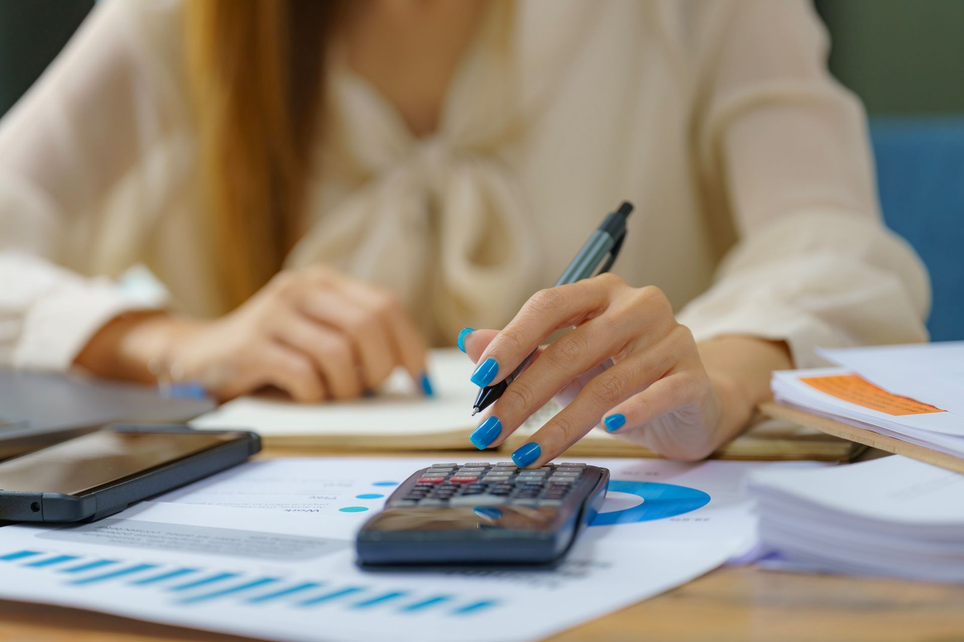 Woman with blue nails using a calculator and pen, working on financial documents at a desk.