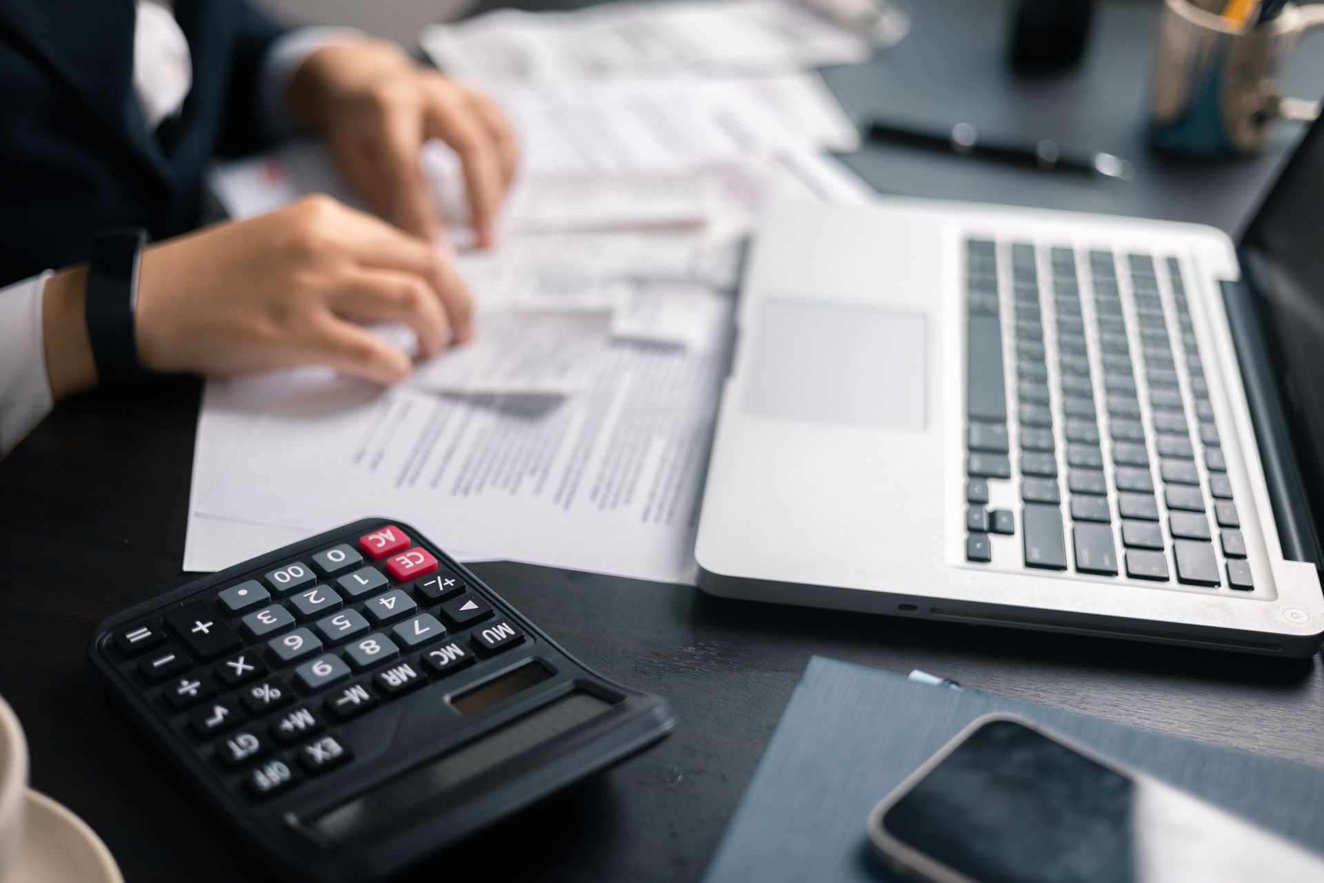 Person reviewing documents at a desk with a laptop, calculator, and phone.