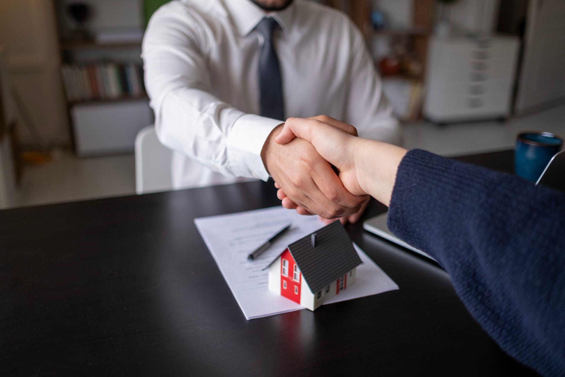 Two people shaking hands over a table with a toy house and documents.