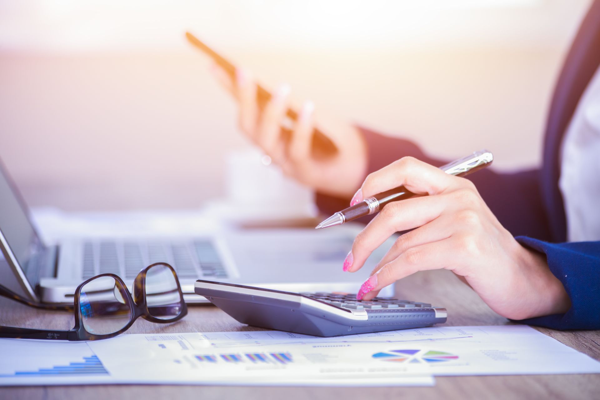 Woman calculating on calculator, holding phone, near laptop and documents.